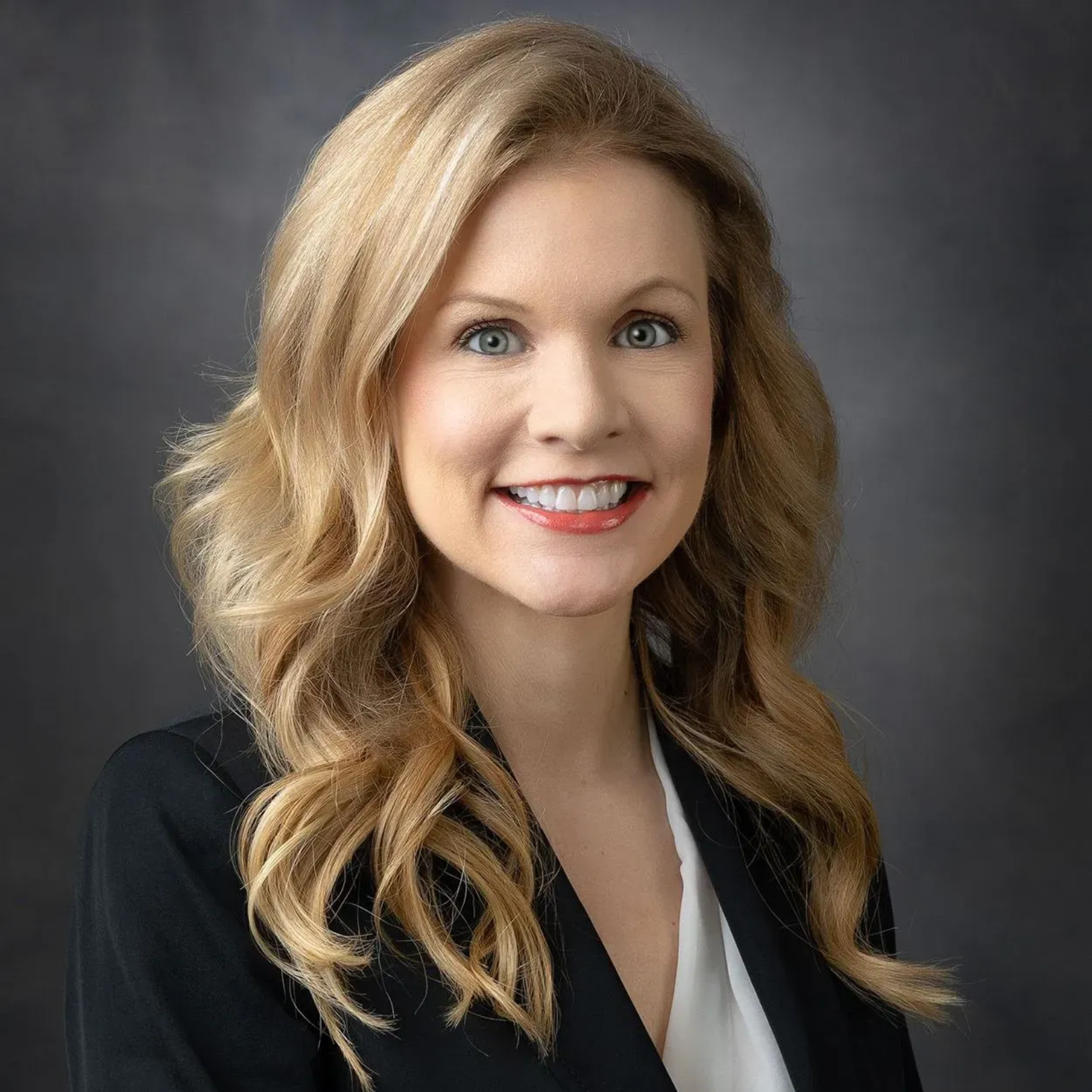 Woman with long blonde hair, smiling, wearing a black blazer over a white shirt, against a dark gray backdrop.