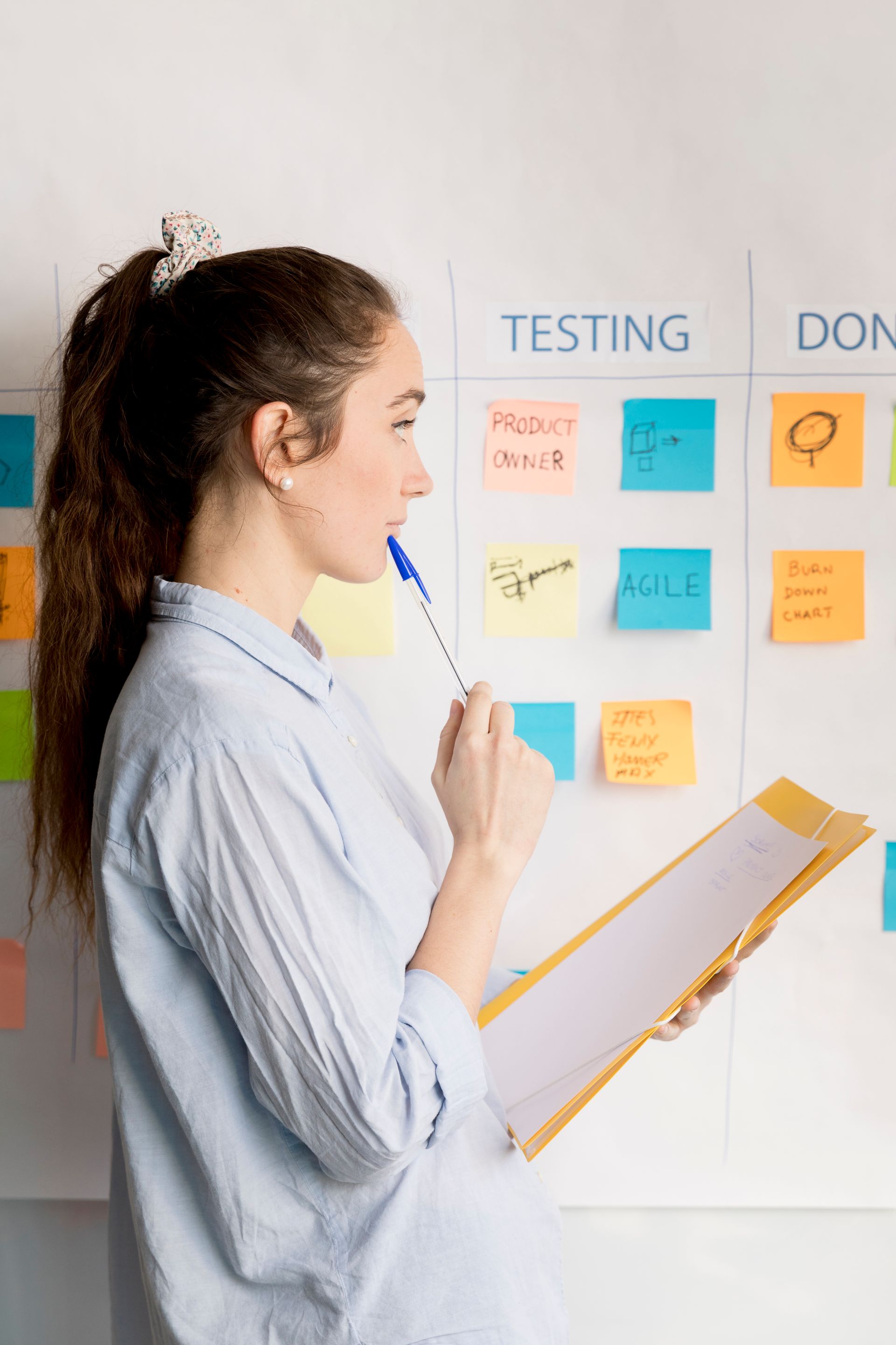 Woman holding pen and clipboard, looking at a Kanban board with sticky notes in a bright office setting.