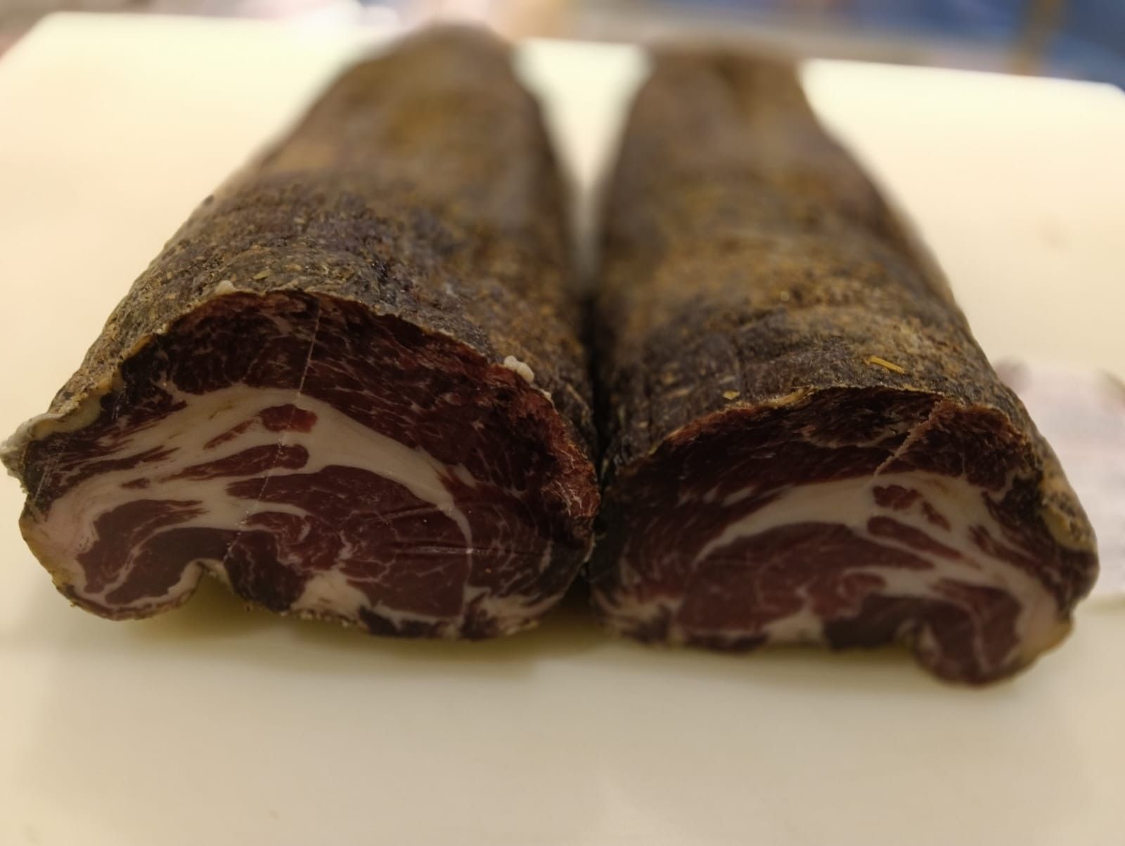 Salumi stagionati appesi su rastrelliere, etichettati con i prezzi, contro una parete piastrellata.