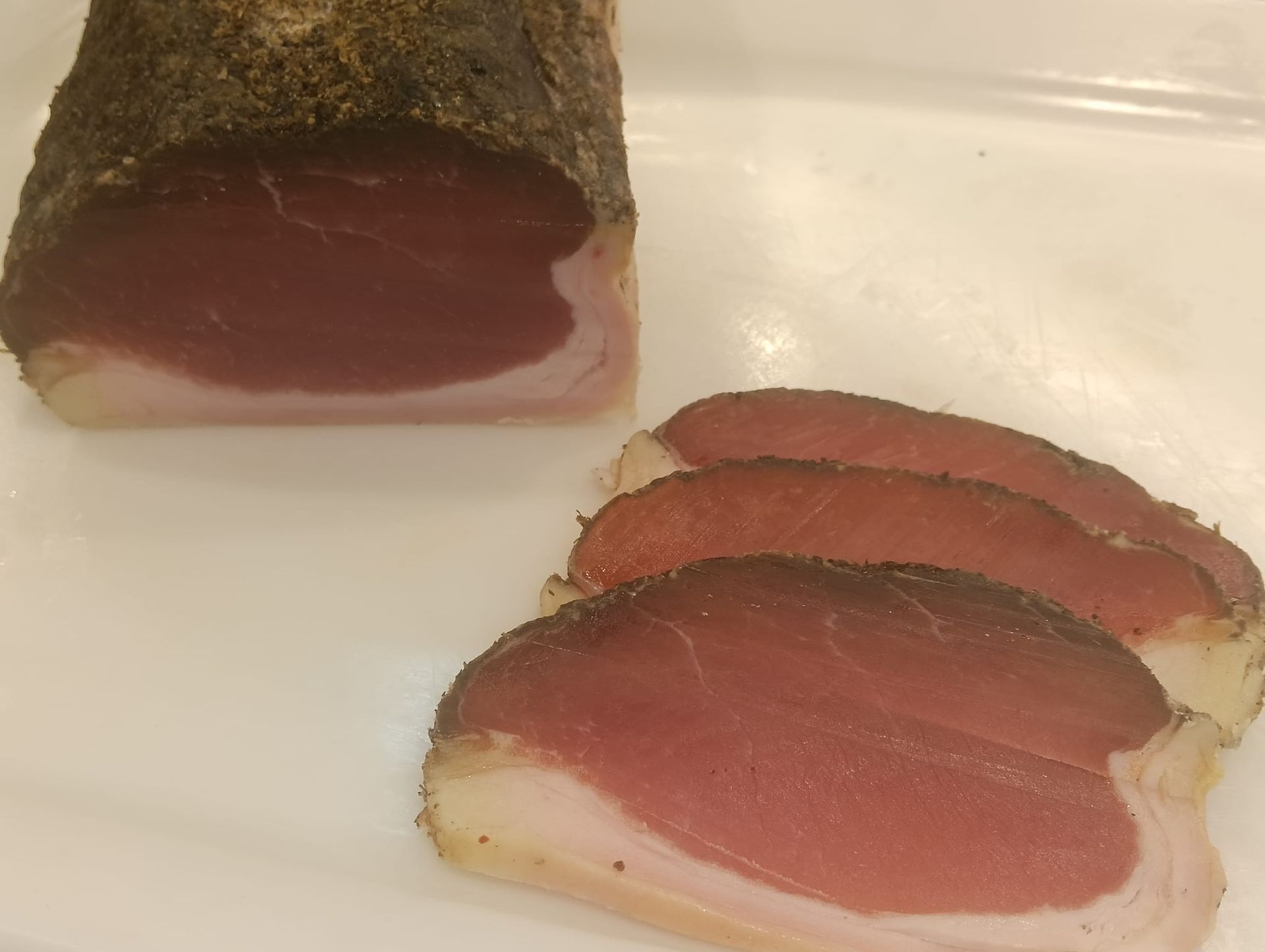 Salumi stagionati appesi su rastrelliere, etichettati con i prezzi, contro una parete piastrellata.