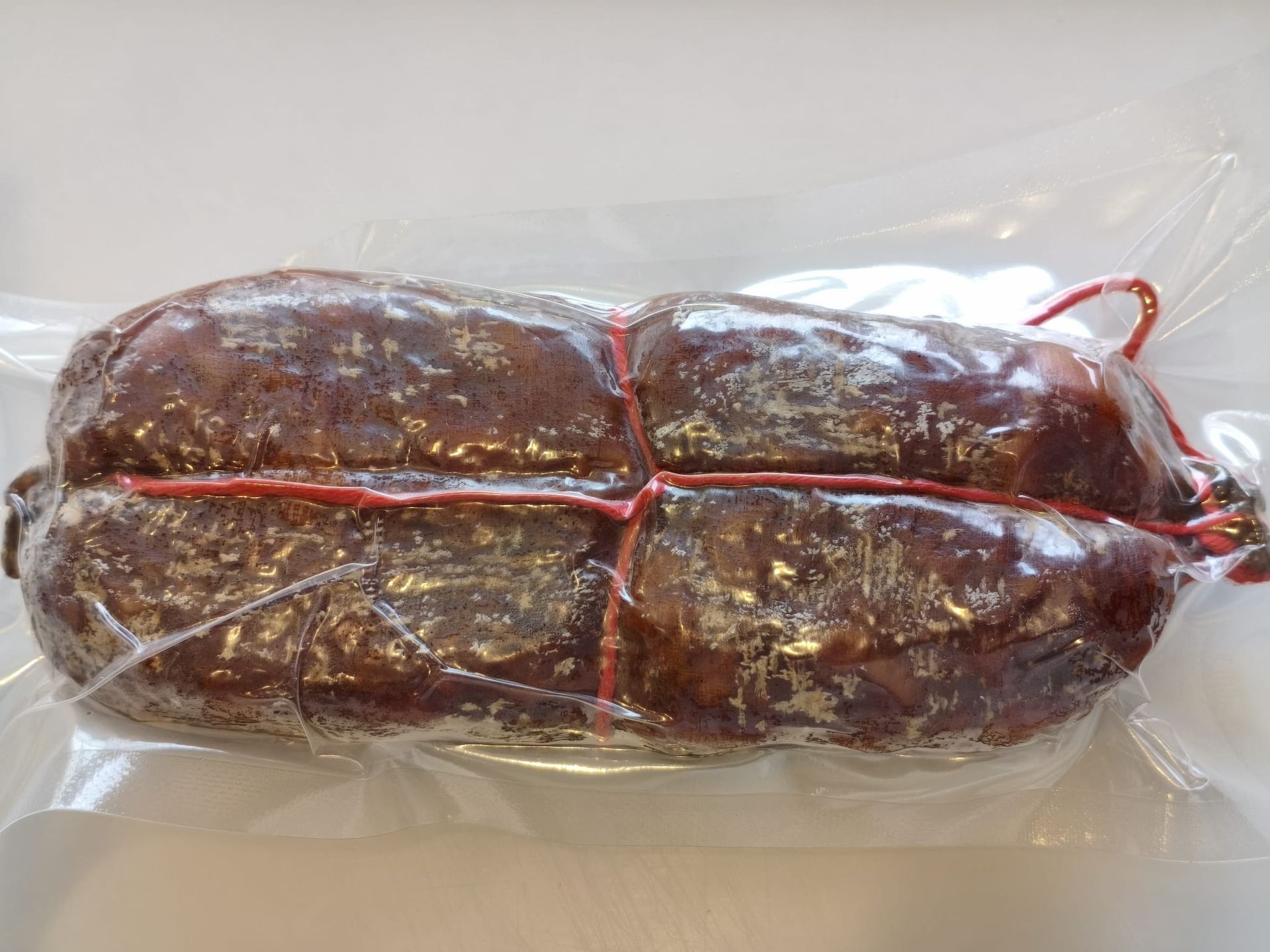 Salumi stagionati appesi su rastrelliere, etichettati con i prezzi, contro una parete piastrellata.