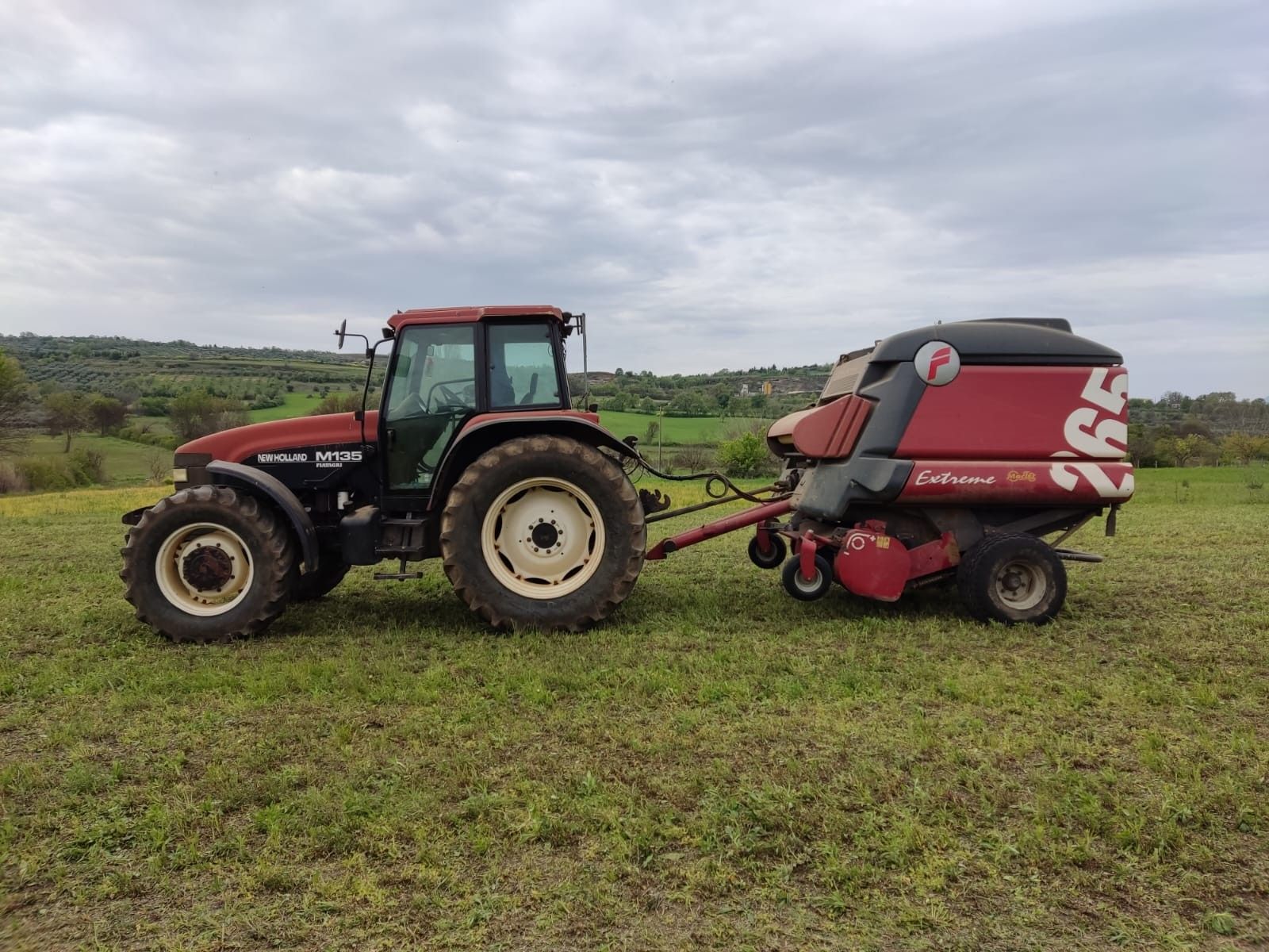 Un trattore rosso traina una rotopressa John Deere in un campo erboso in una giornata nuvolosa.