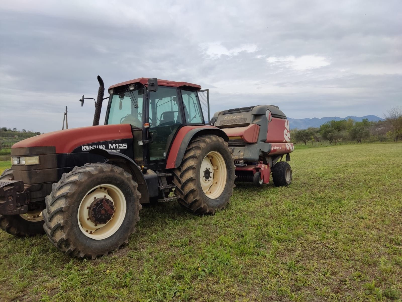Un trattore rosso traina una pressa per fieno in un campo erboso sotto un cielo nuvoloso.