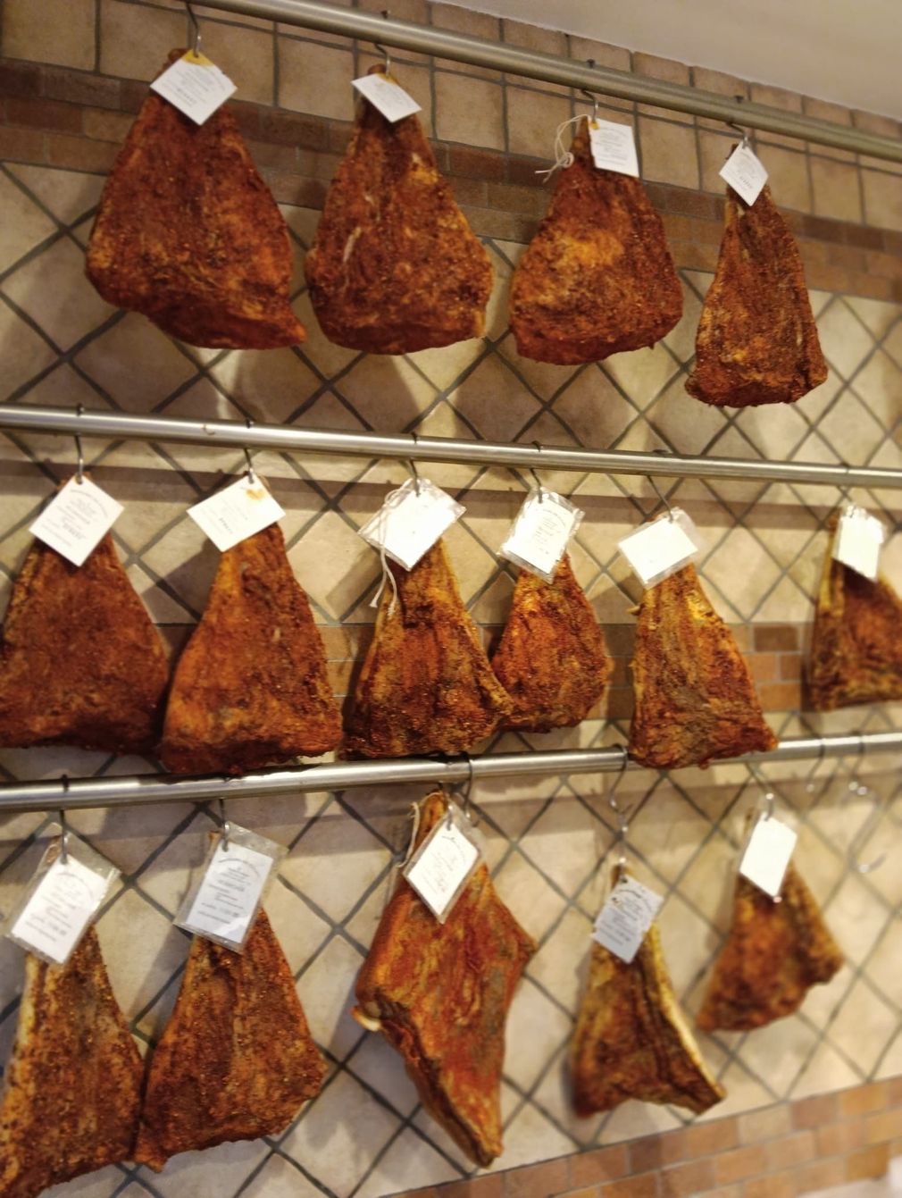 Salumi stagionati appesi su rastrelliere, etichettati con i prezzi, contro una parete piastrellata.