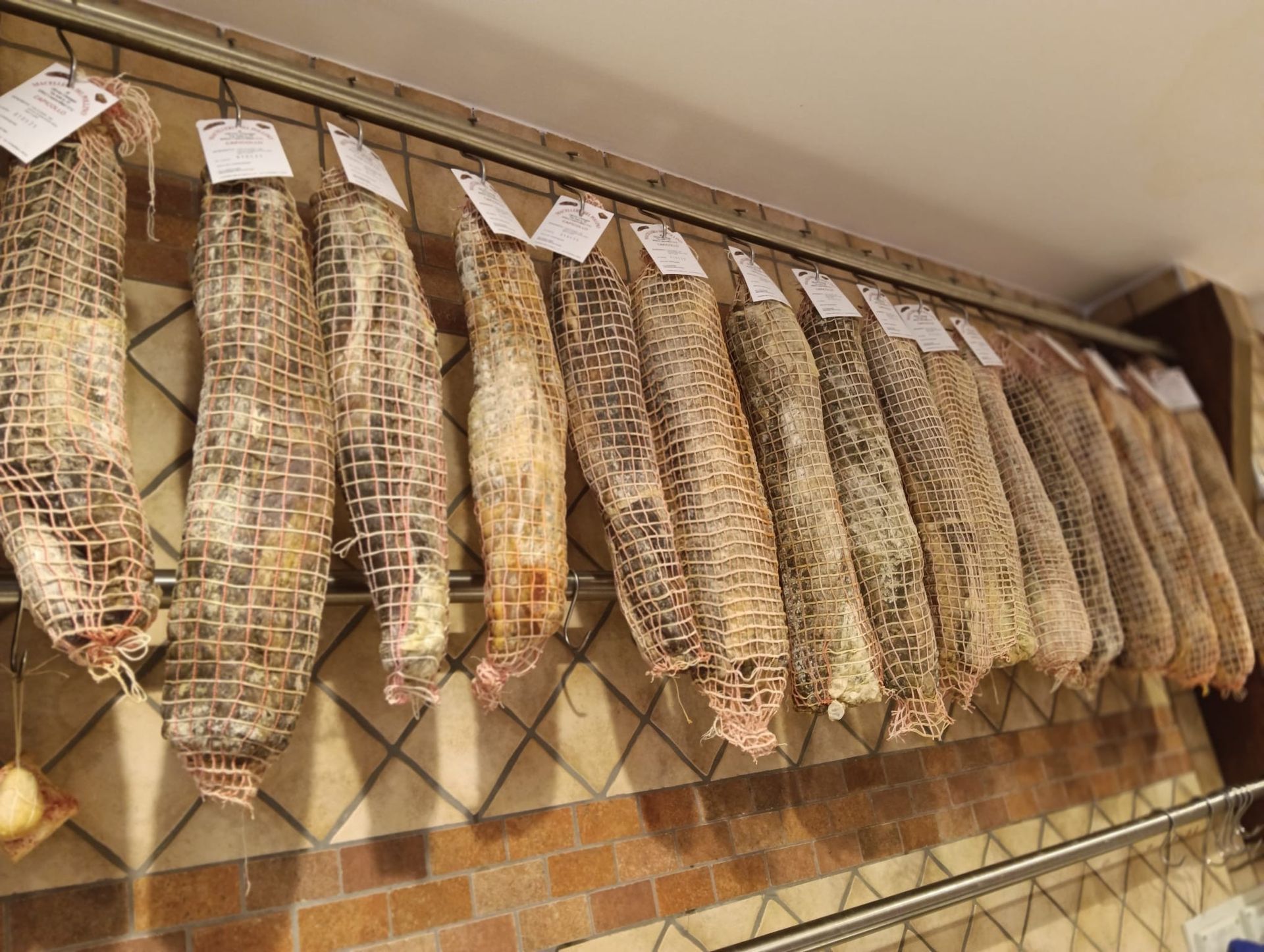 Fila di salumi appesi in un negozio, avvolti in una rete e fissati a una rastrelliera di metallo.