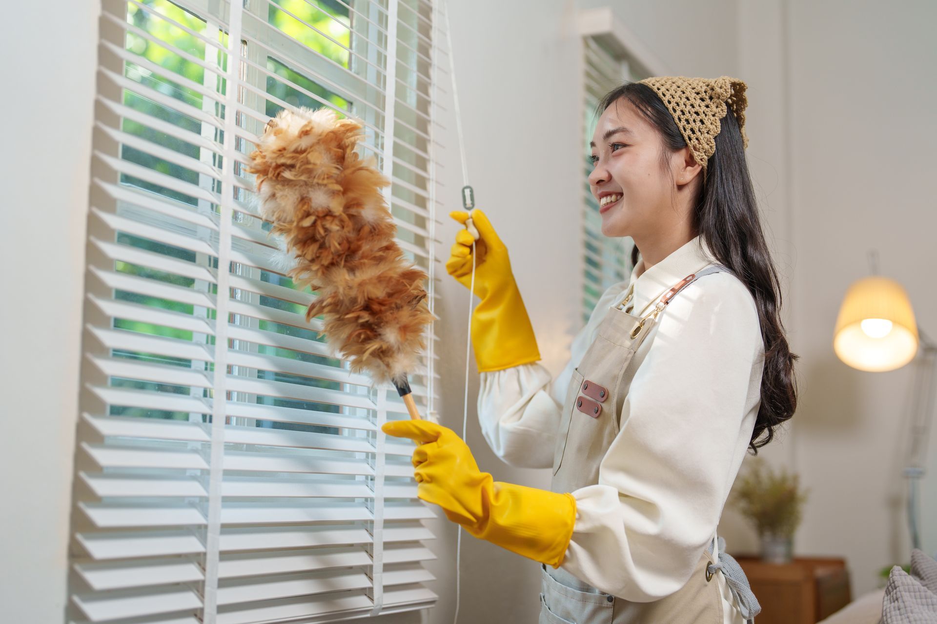 A woman in yellow gloves is cleaning blinds with a duster.