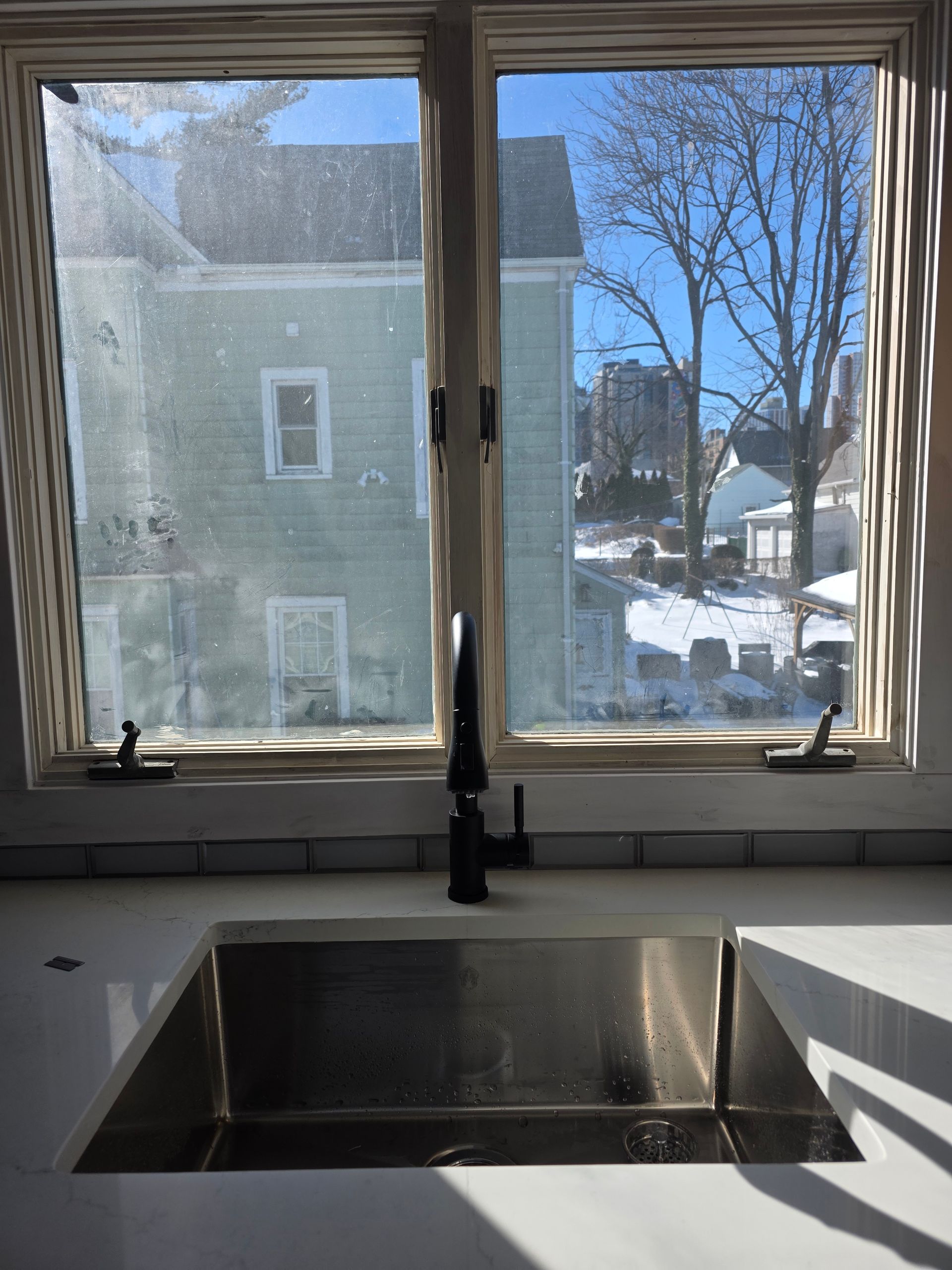 Stainless steel kitchen sink under a window with a view of a snowy neighborhood on a sunny day.