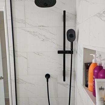 Black shower fixtures against white marble tile in a bathroom. A recessed shelf holds toiletries.
