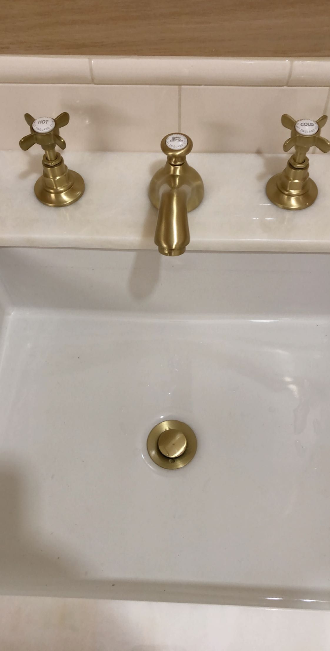 White bathroom sink with gold faucets and drain, set against a light-colored tile backsplash.