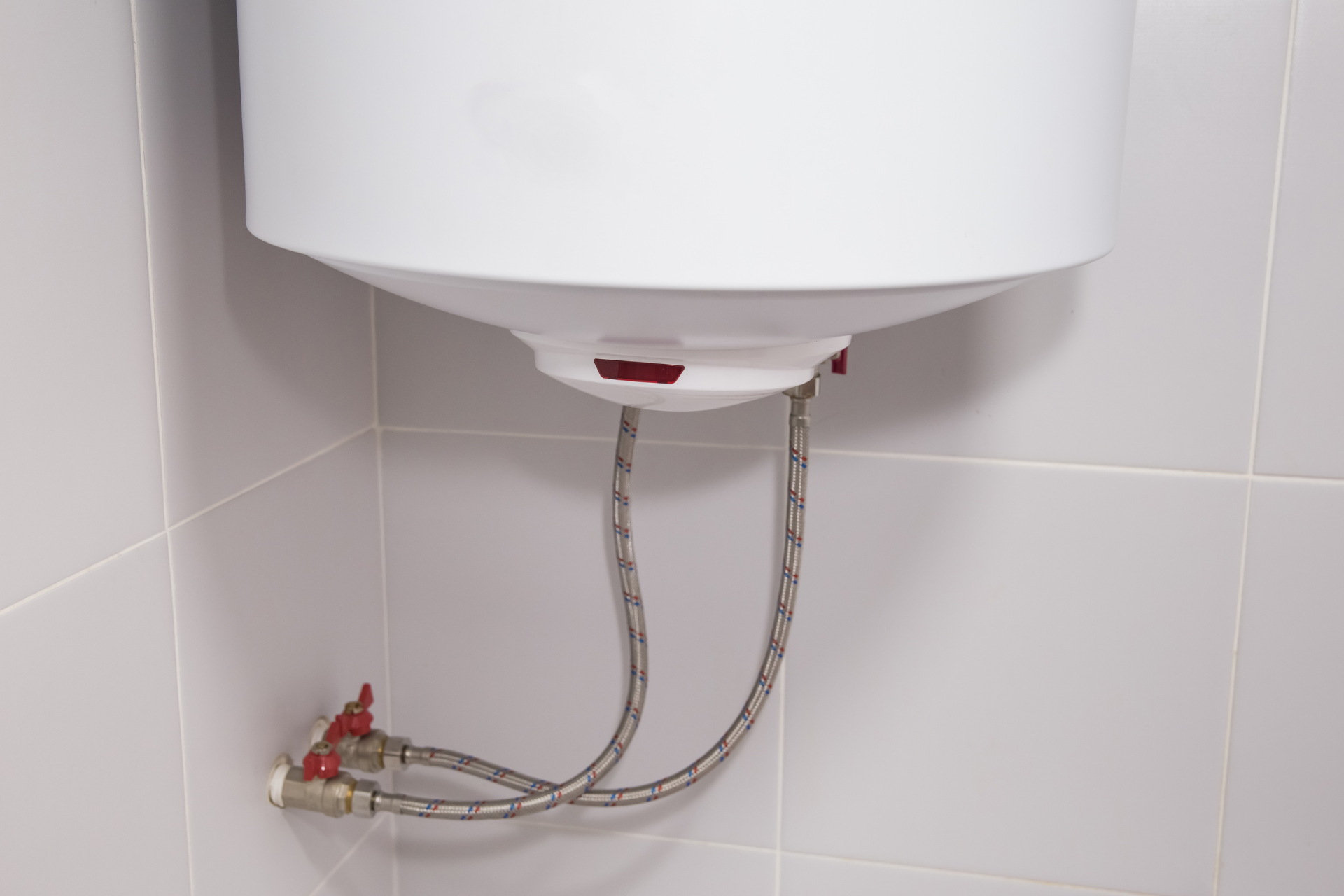 White water heater mounted on tiled wall, with flexible braided hoses connected to it.