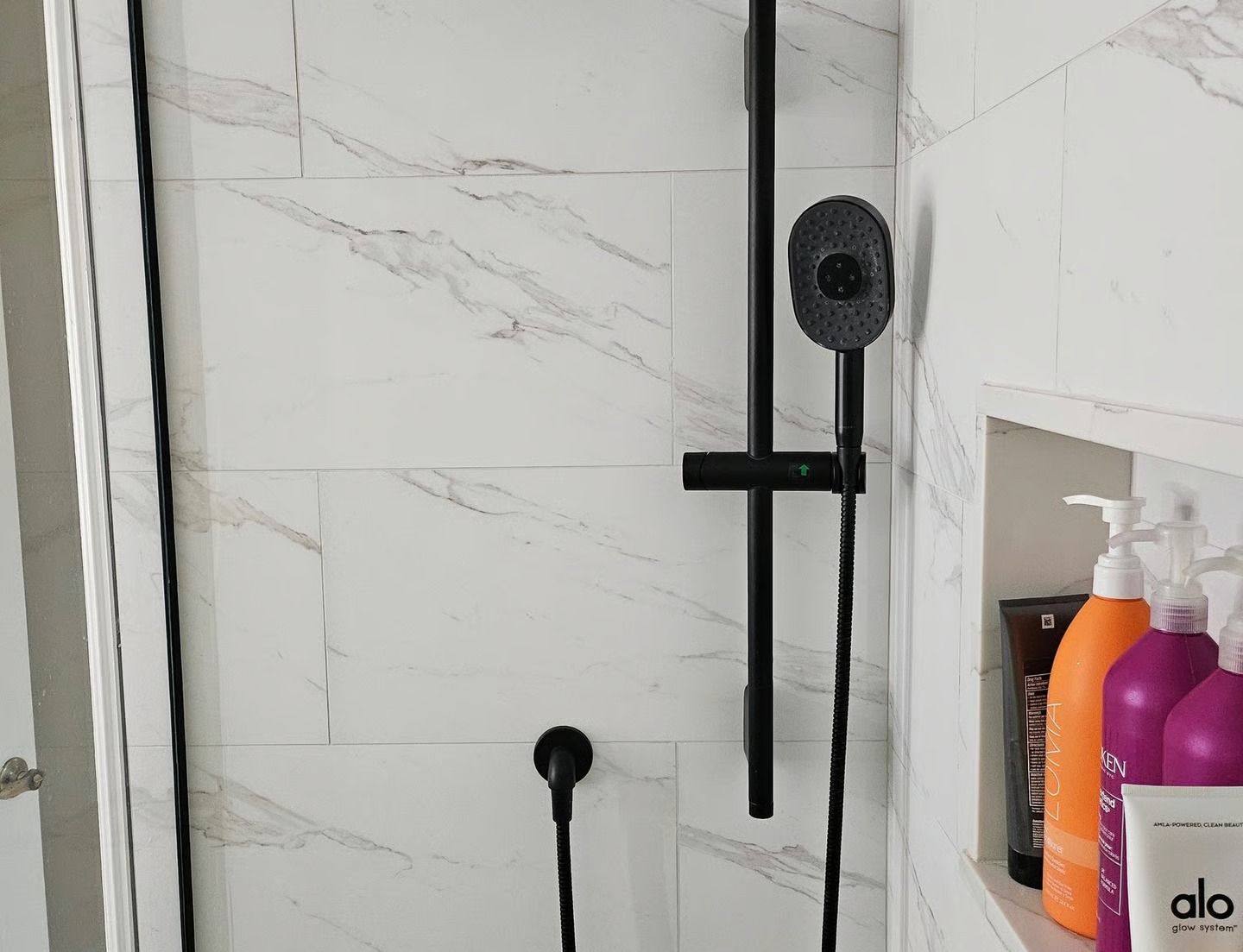 Black showerhead and fixtures against white marble-patterned tile wall. Recessed shelf with toiletries.
