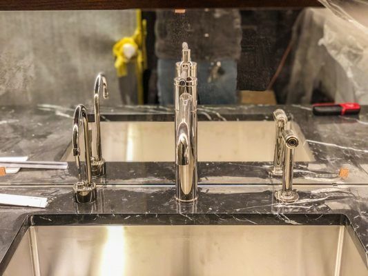 Three chrome faucets installed on a black marble countertop above stainless steel sinks, with a person in the background.