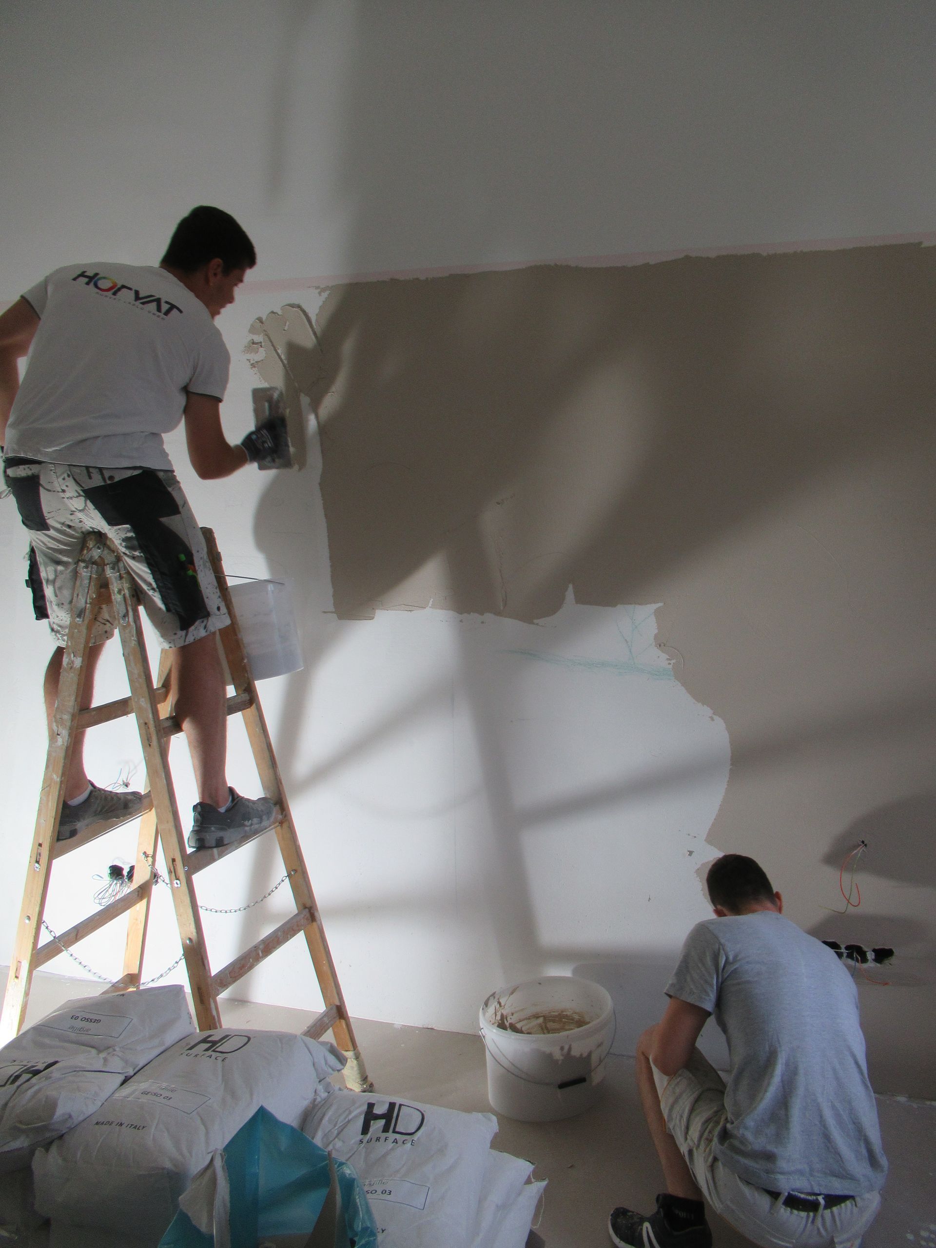 Zwei Männer streichen eine Wand und einer sitzt auf einer Leiter