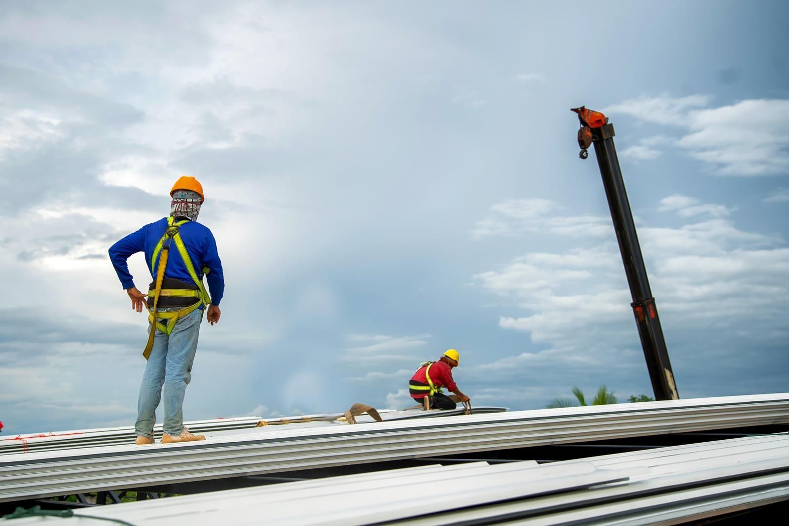 Construction workers wearing safety harnesses and hard hats installing metal roofing sheets under a cloudy sky with a crane.