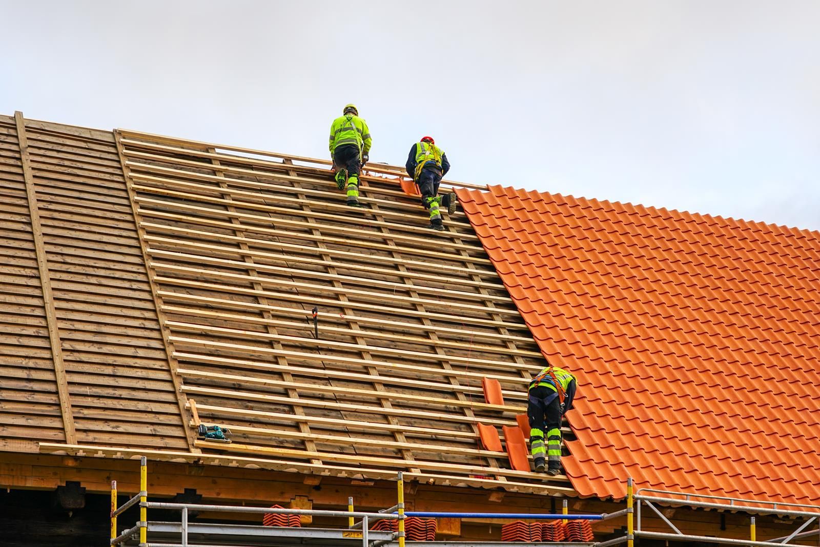 Three workers in safety gear installing orange roof tiles on a wooden roof frame, using scaffolding for support.