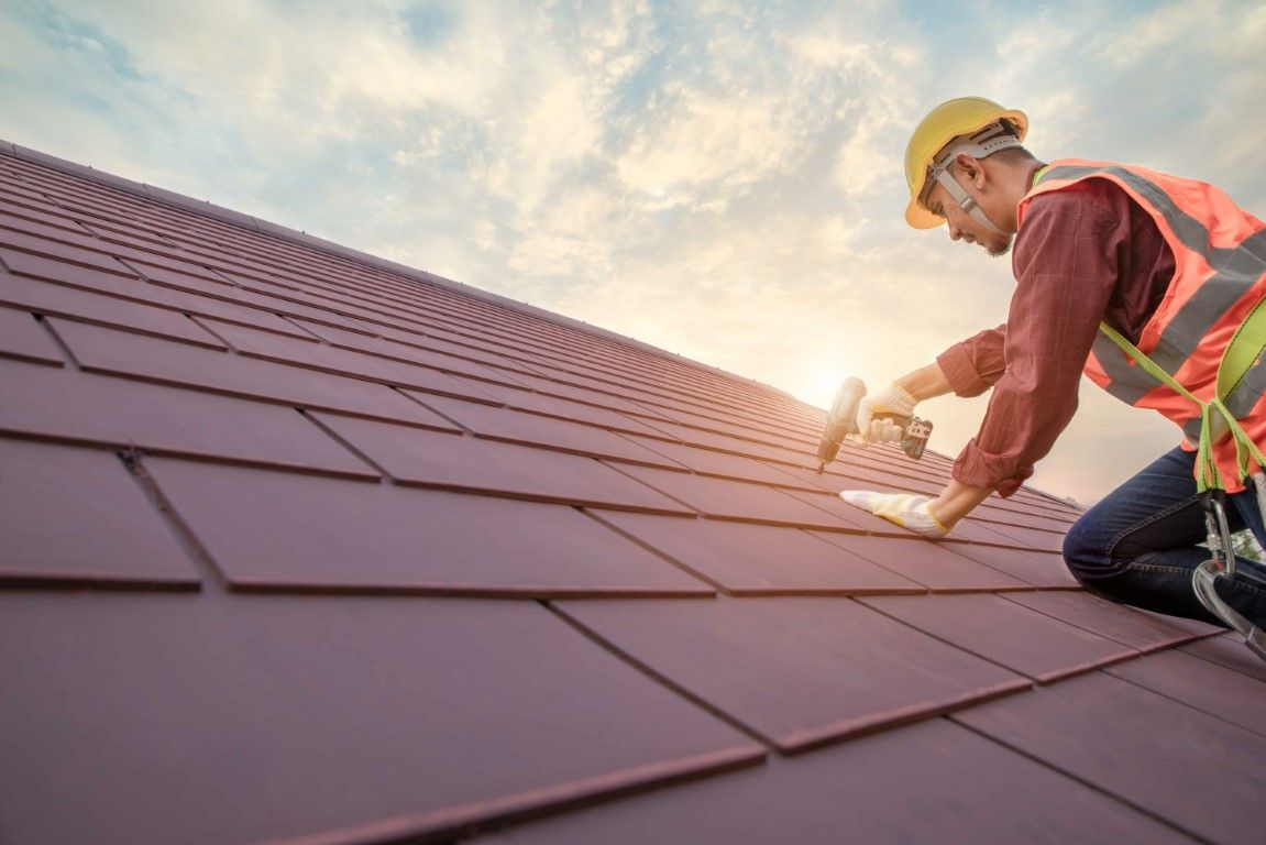 Construction worker installing red roof shingles with a drill while wearing safety gear under a cloudy sunset sky.