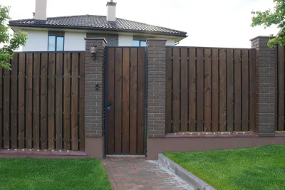 Wooden fence with gate, brick pillars, and house in the background. Green lawn and brick pathway in front.