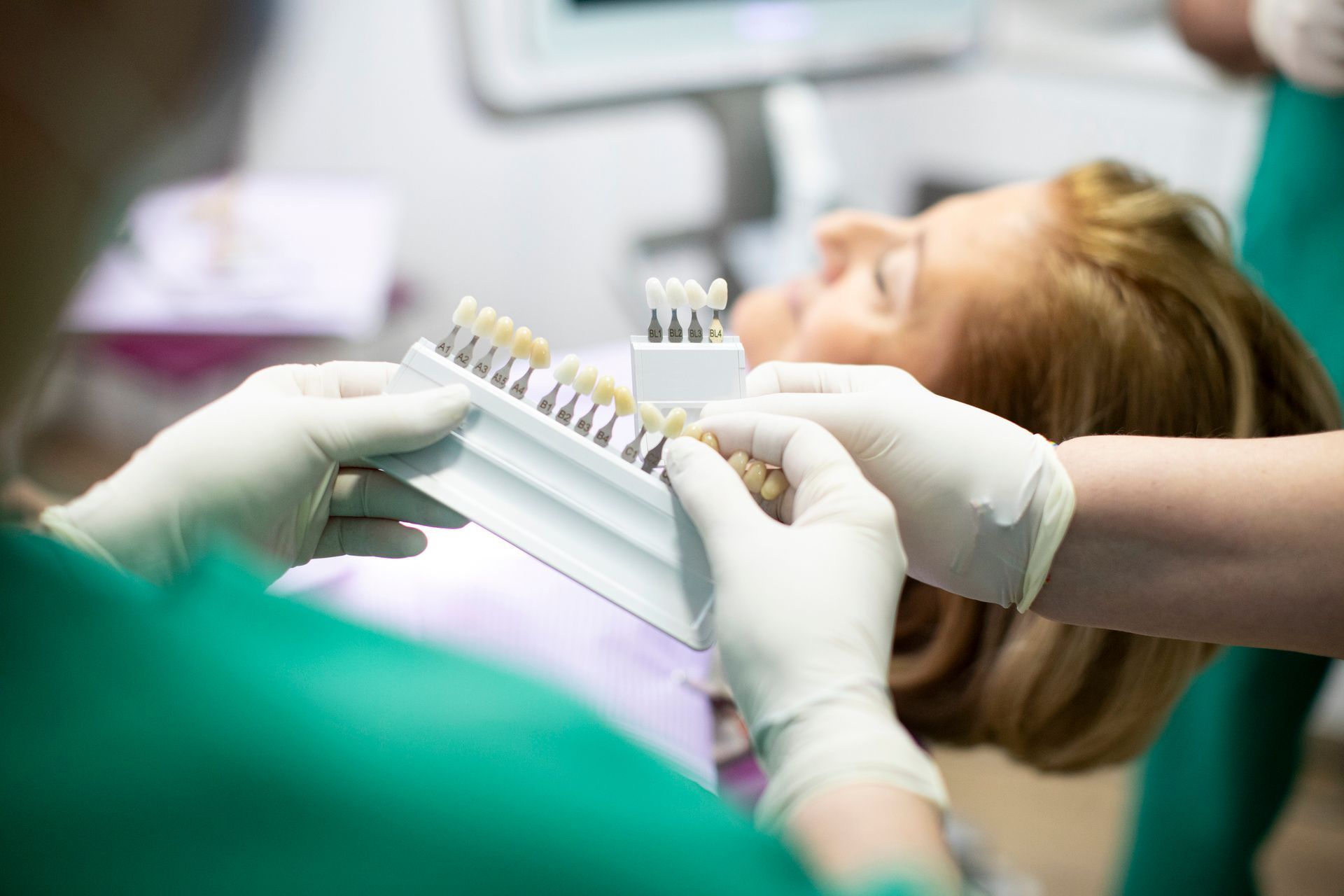Dentist holding tooth shade guide to match dental crowns for female patient in chair.