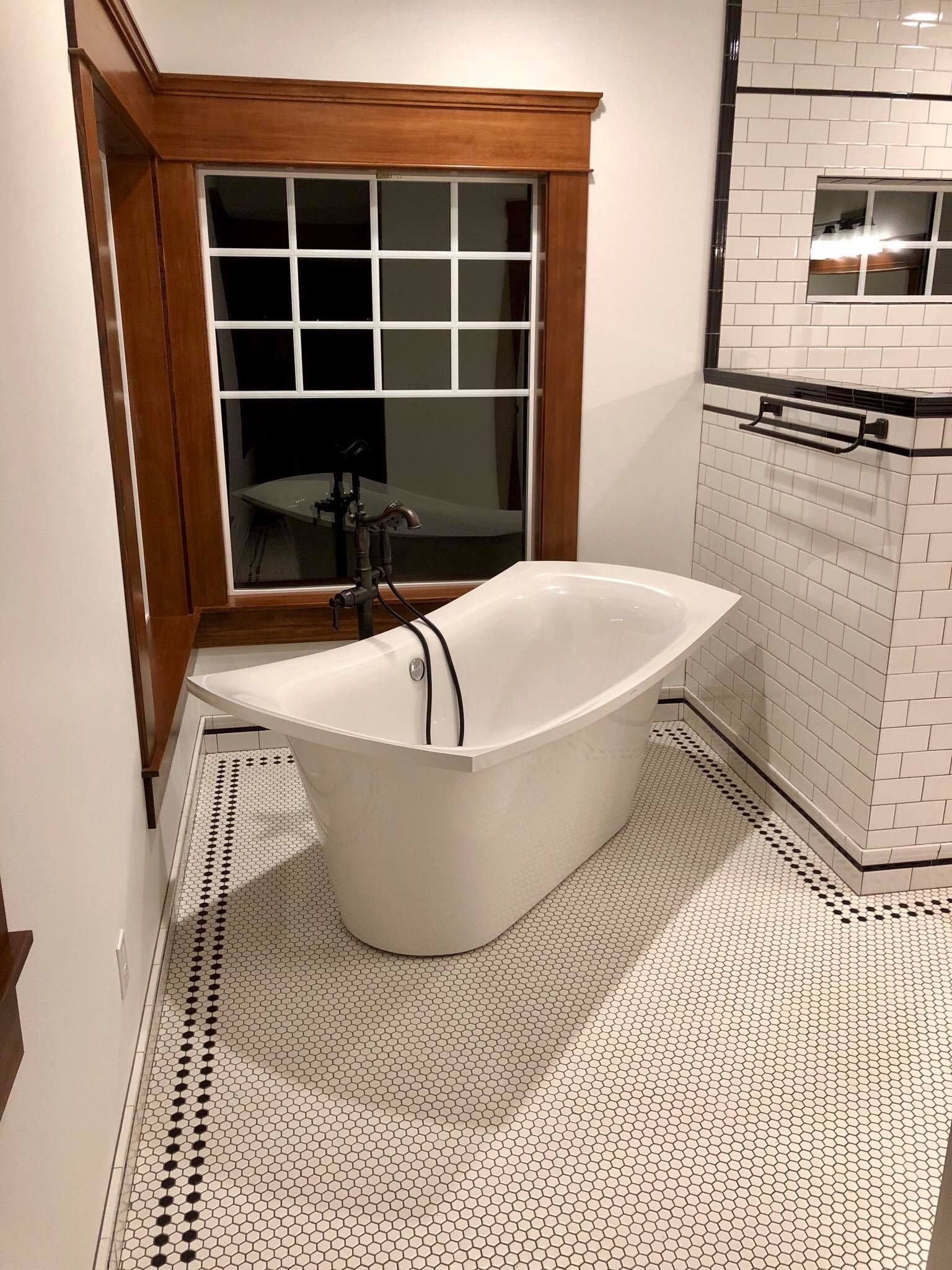 A white claw-foot bathtub under a window in a bathroom with patterned floor tiles.