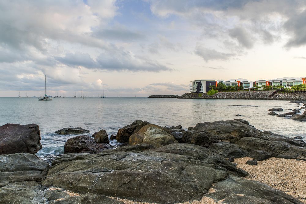 A Rocky Beach With a Boat in the Water and Buildings in the Background — Hair By Jell In Cannonvale, QLD