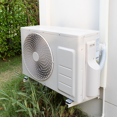 Outdoor wall-mounted air conditioner unit beside a white building, with greenery nearby