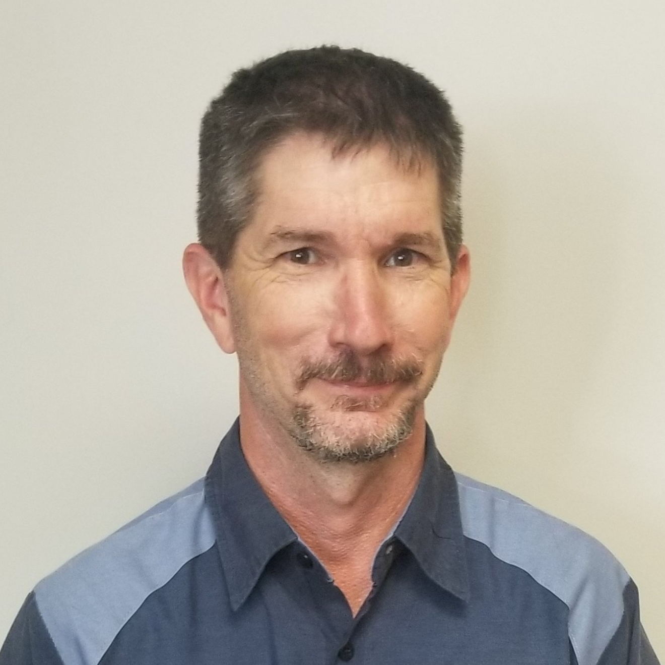 A man with a beard is wearing a blue and grey shirt.