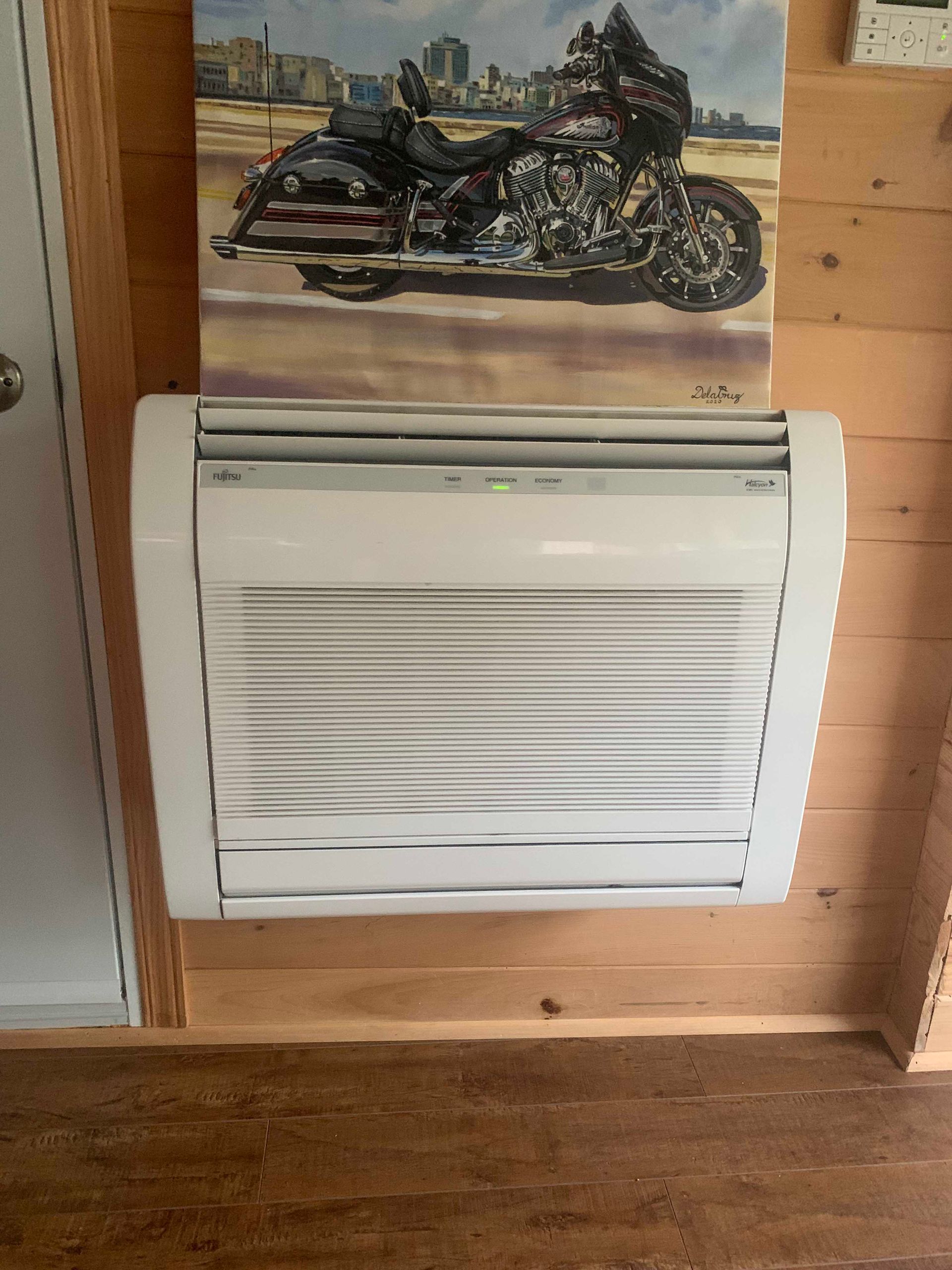 Unité de climatisation murale blanche sous un tableau de moto sur un mur lambrissé.