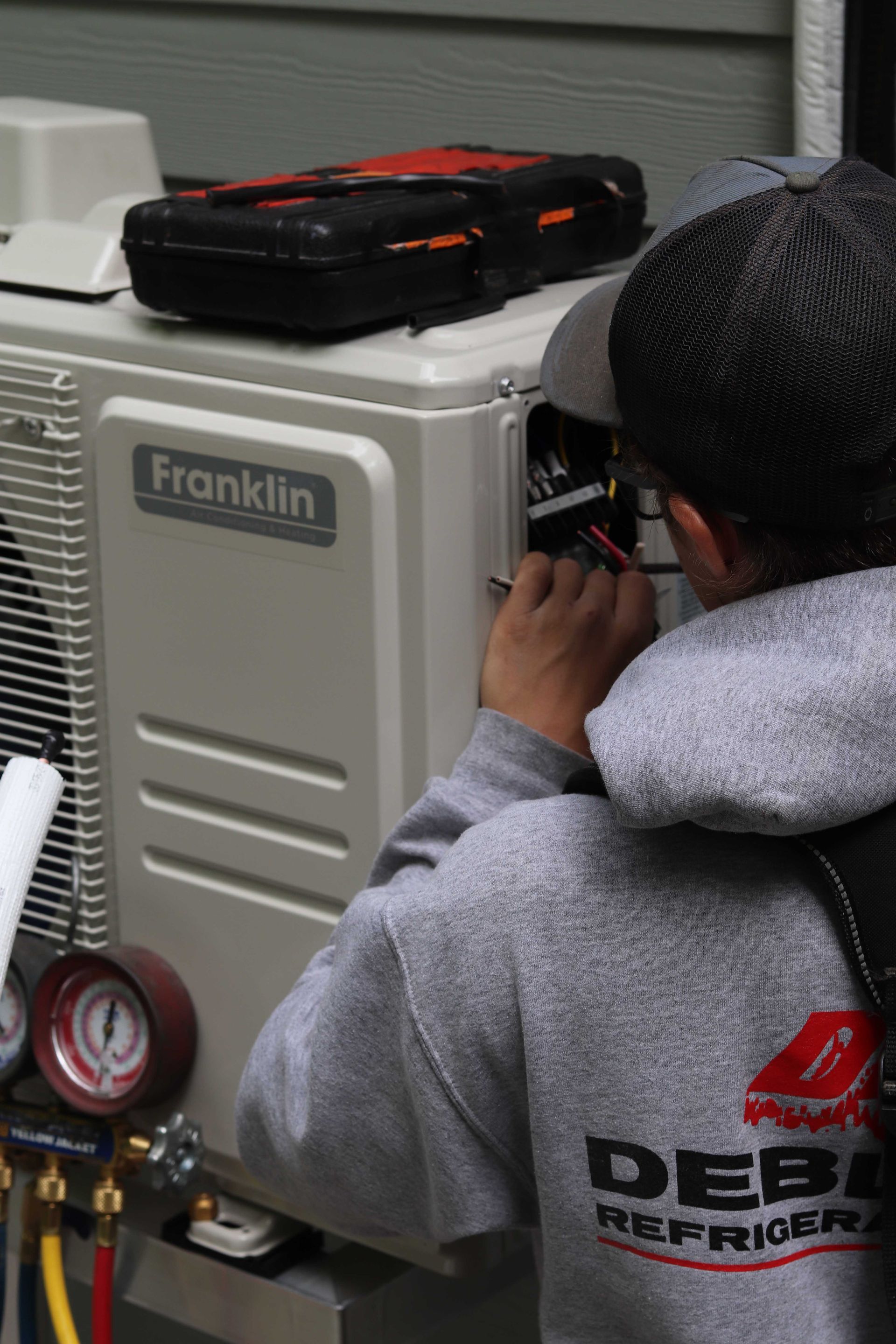 Technicien CVC en sweat-shirt gris travaillant sur une unité de climatisation Franklin à l'extérieur, une boîte à outils posée dessus.