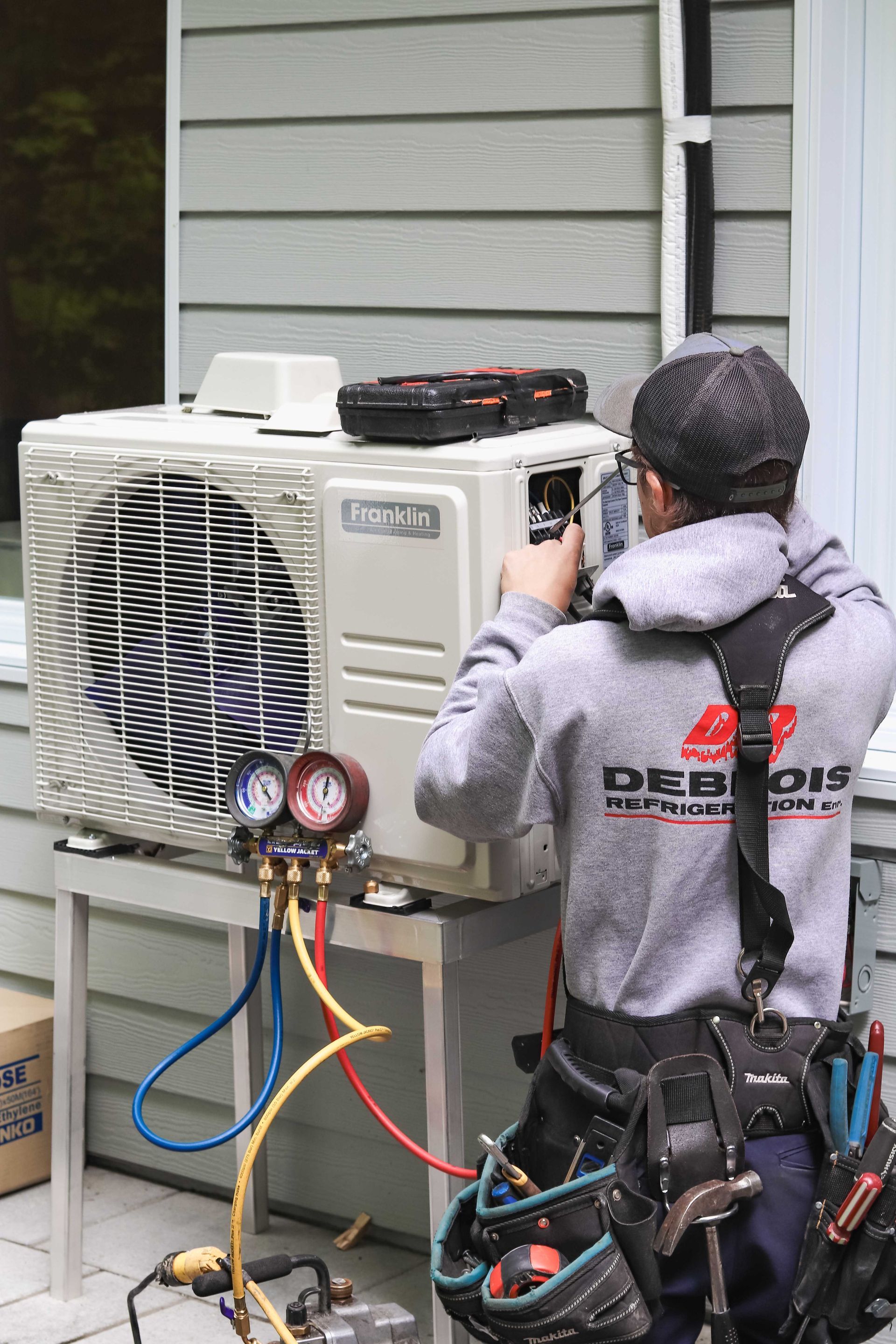 Technicien CVC travaillant sur une unité de climatisation extérieure, à l'aide de manomètres. Bâtiment gris en arrière-plan.