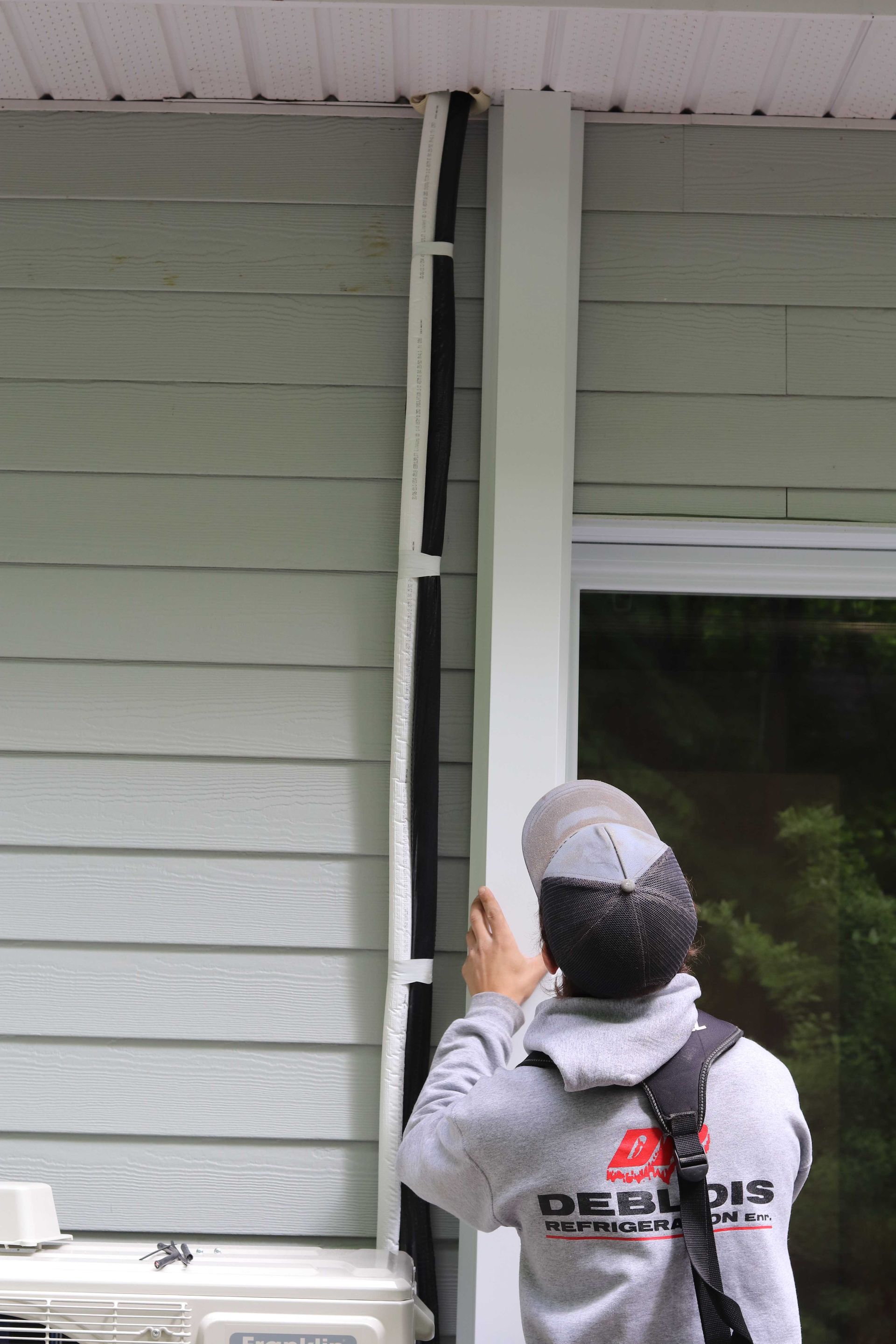 Une personne installe des tubes isolés sur le côté d'un bâtiment, près d'une fenêtre et d'un climatiseur.