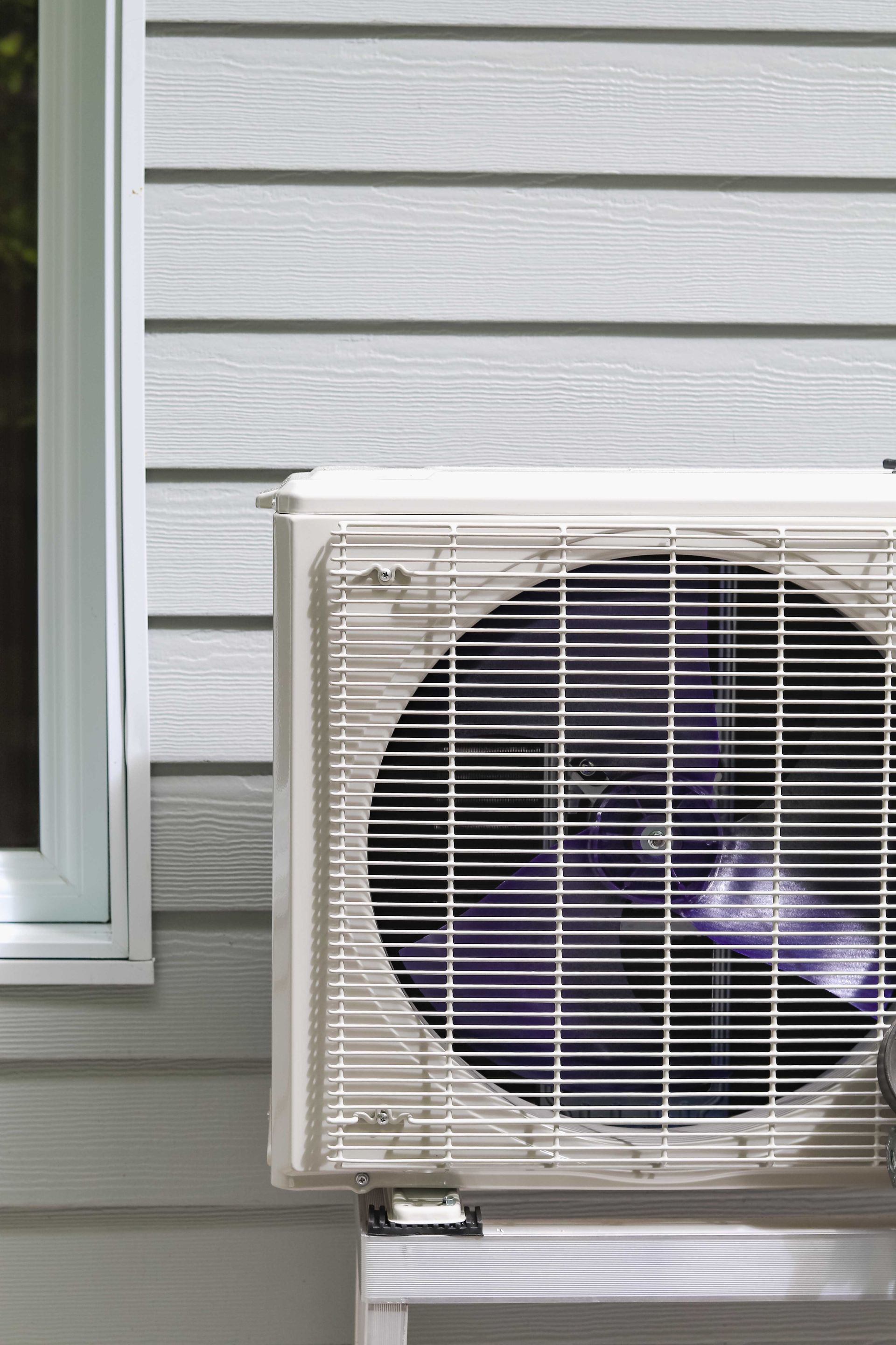 Unité de climatisation extérieure fixée sur un mur bardé gris clair, à côté d'une fenêtre.