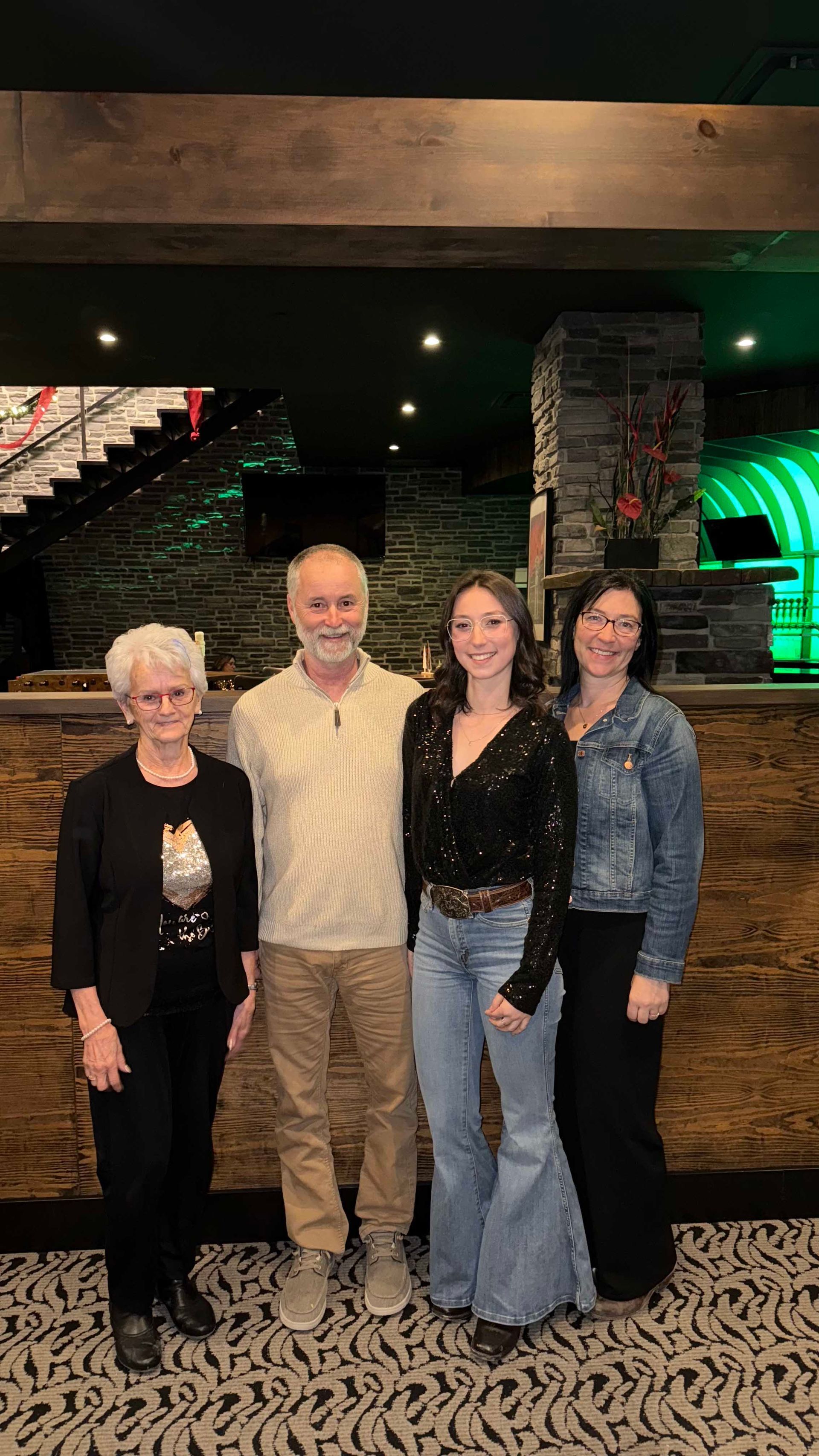 Quatre personnes se tiennent ensemble dans une salle. Deux femmes et un homme. L'une d'elles, vêtue d'un jean et d'un haut noir à paillettes, sourit. Intérieur sombre.