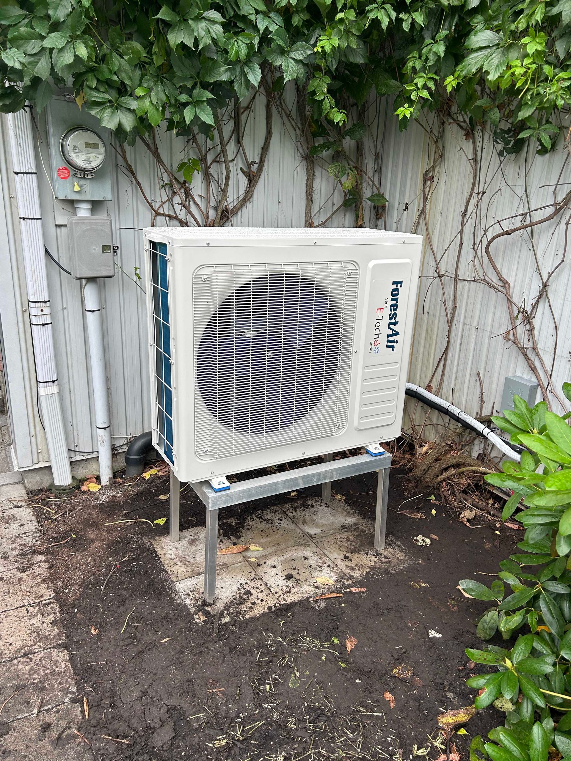 Unité de climatisation sur un support métallique, contre une clôture végétalisée.