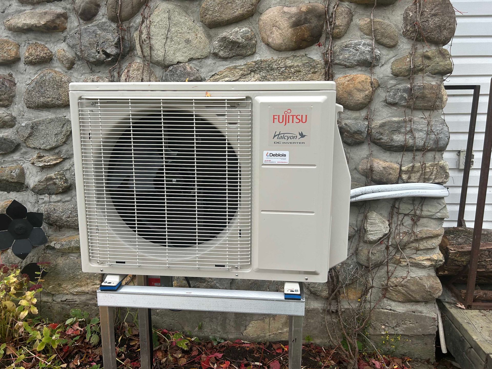 Unité de pompe à chaleur Fujitsu montée sur un support métallique, contre un mur en pierre, en extérieur.