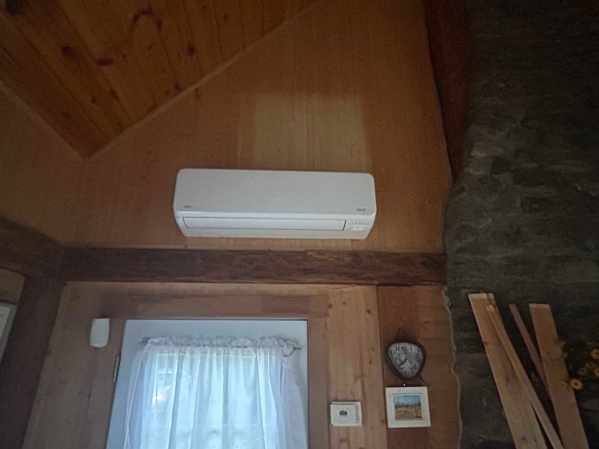 Unité de climatisation blanche fixée sur un mur lambrissé au-dessus d'une fenêtre munie d'un rideau blanc.