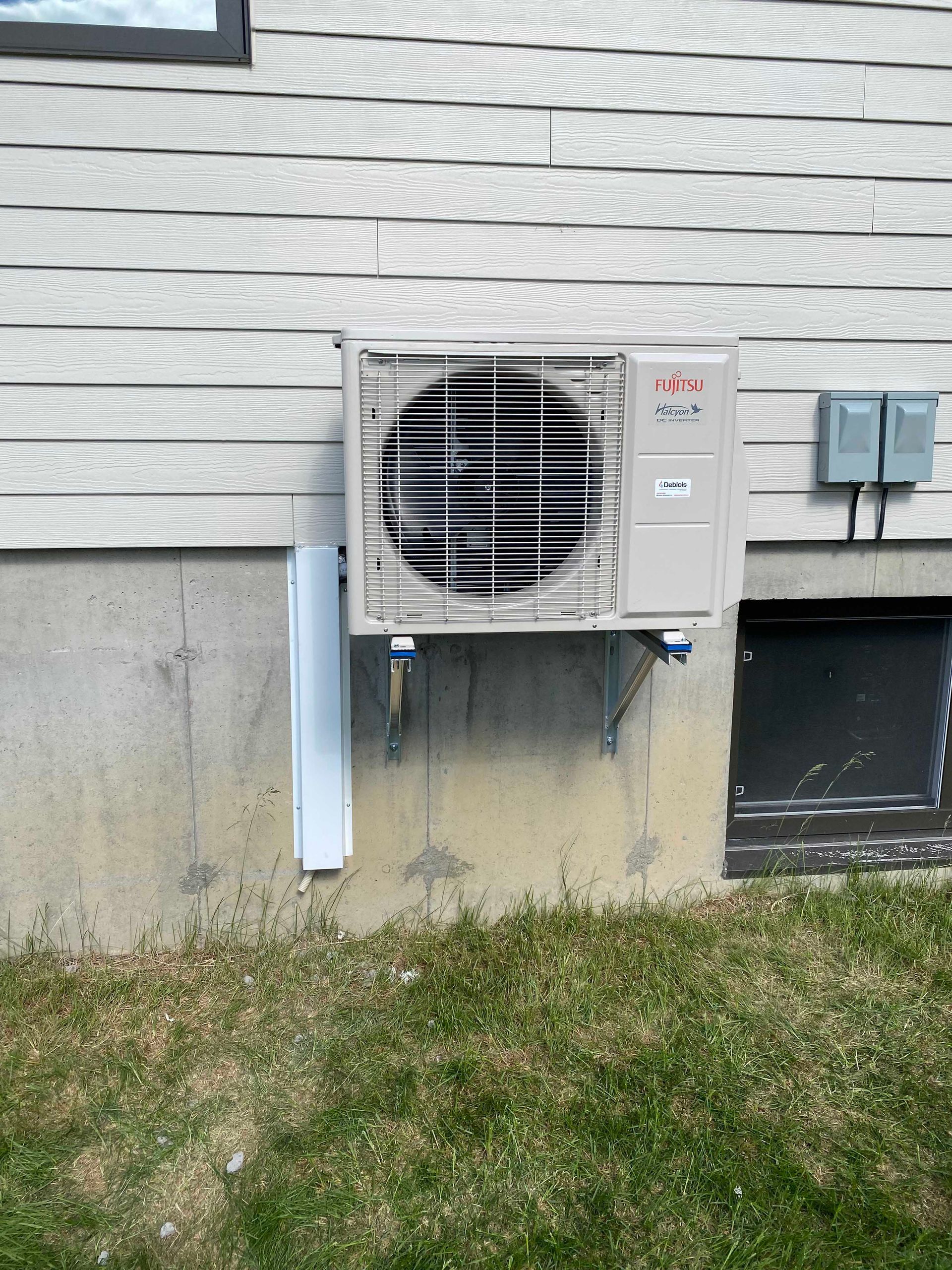 Unité de climatisation extérieure montée sur un mur en béton, avec un revêtement extérieur au-dessus et de l'herbe en dessous.