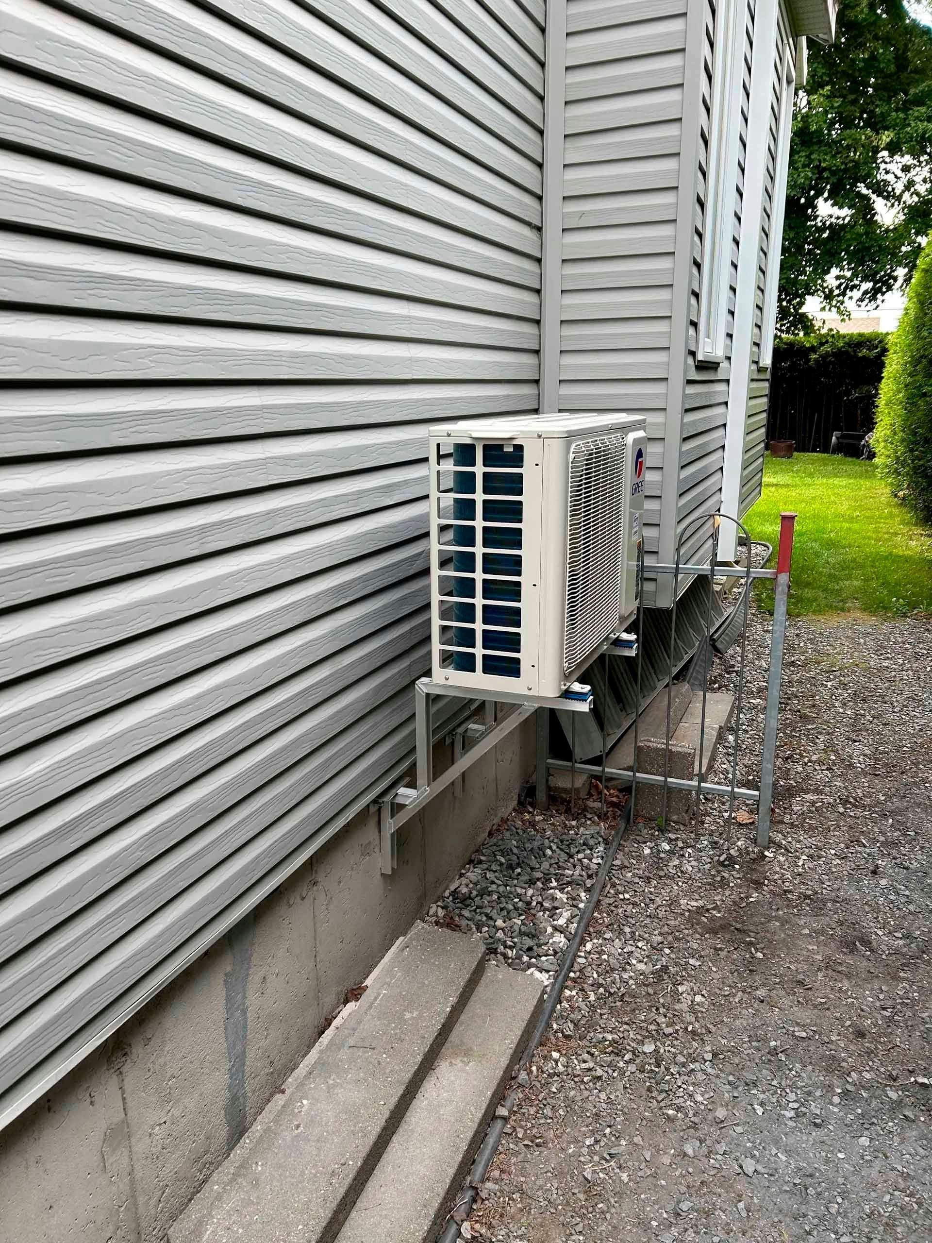Unité de climatisation montée sur les fondations d'un bâtiment à bardage gris, sur un sol en gravier.