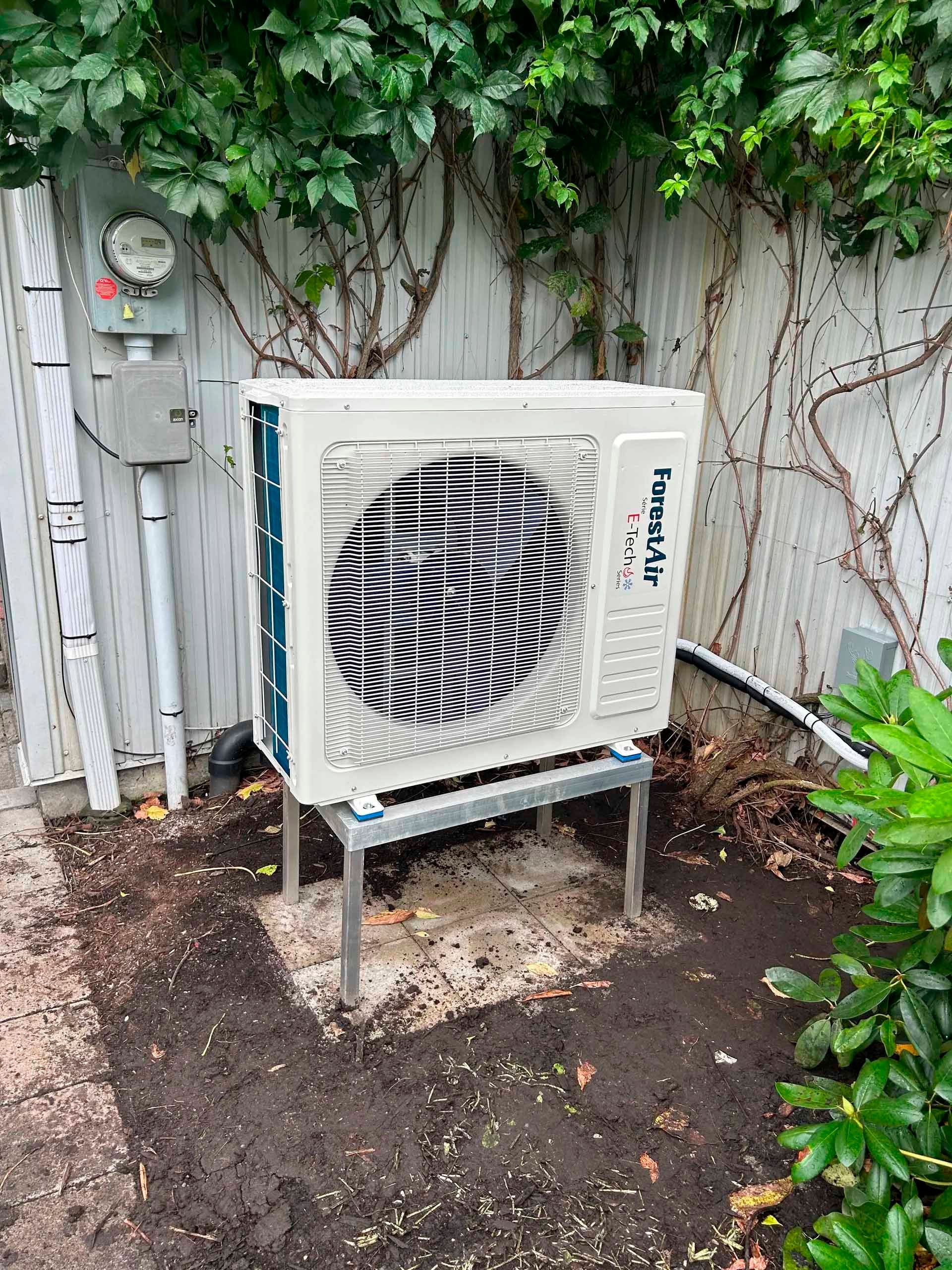 Un climatiseur sur pieds métalliques, adossé à une clôture blanche recouverte de lierre.