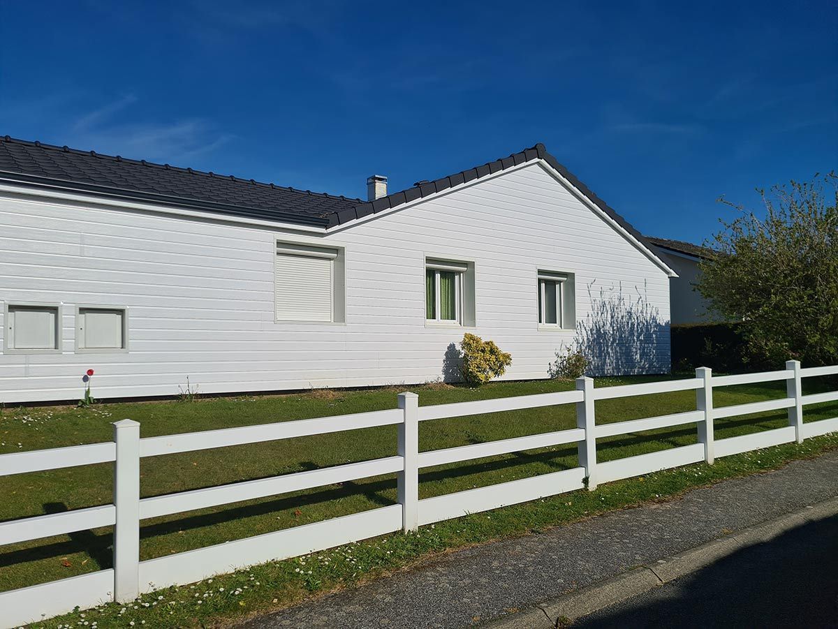 Une maison blanche au toit sombre, derrière une clôture blanche, se détachant sur un ciel bleu azur 