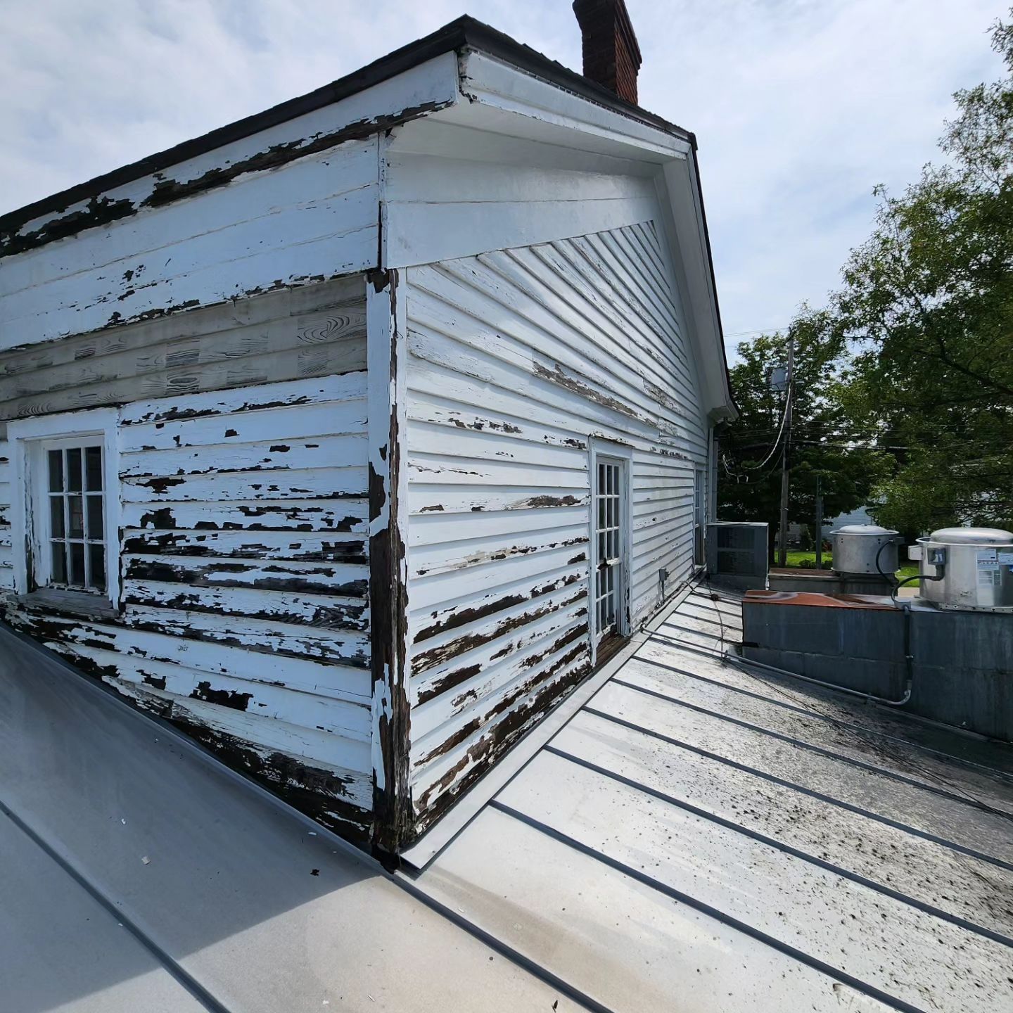 White building with peeling paint on a metal roof, with two windows and a brick chimney.