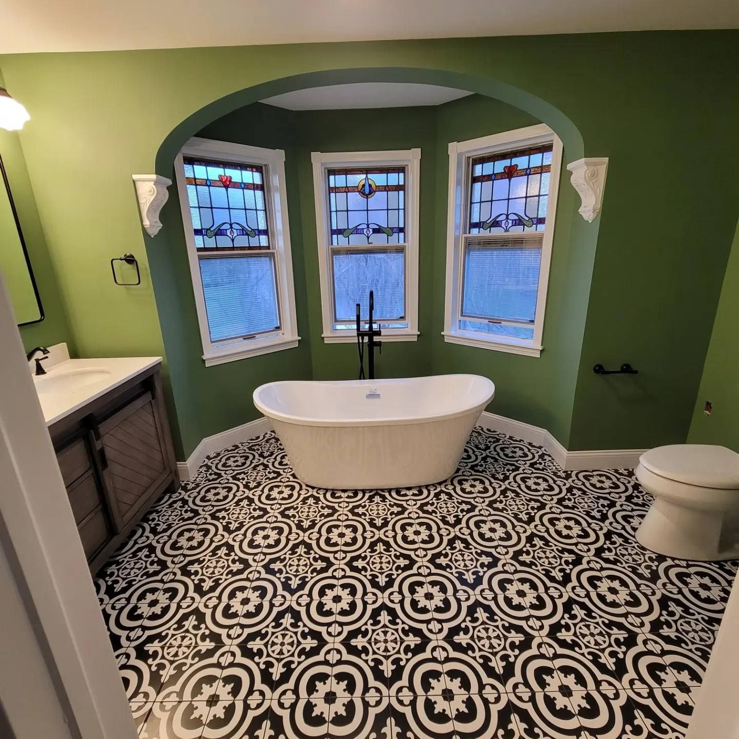 Bathroom with green walls, patterned floor, and a white bathtub beneath arched windows.