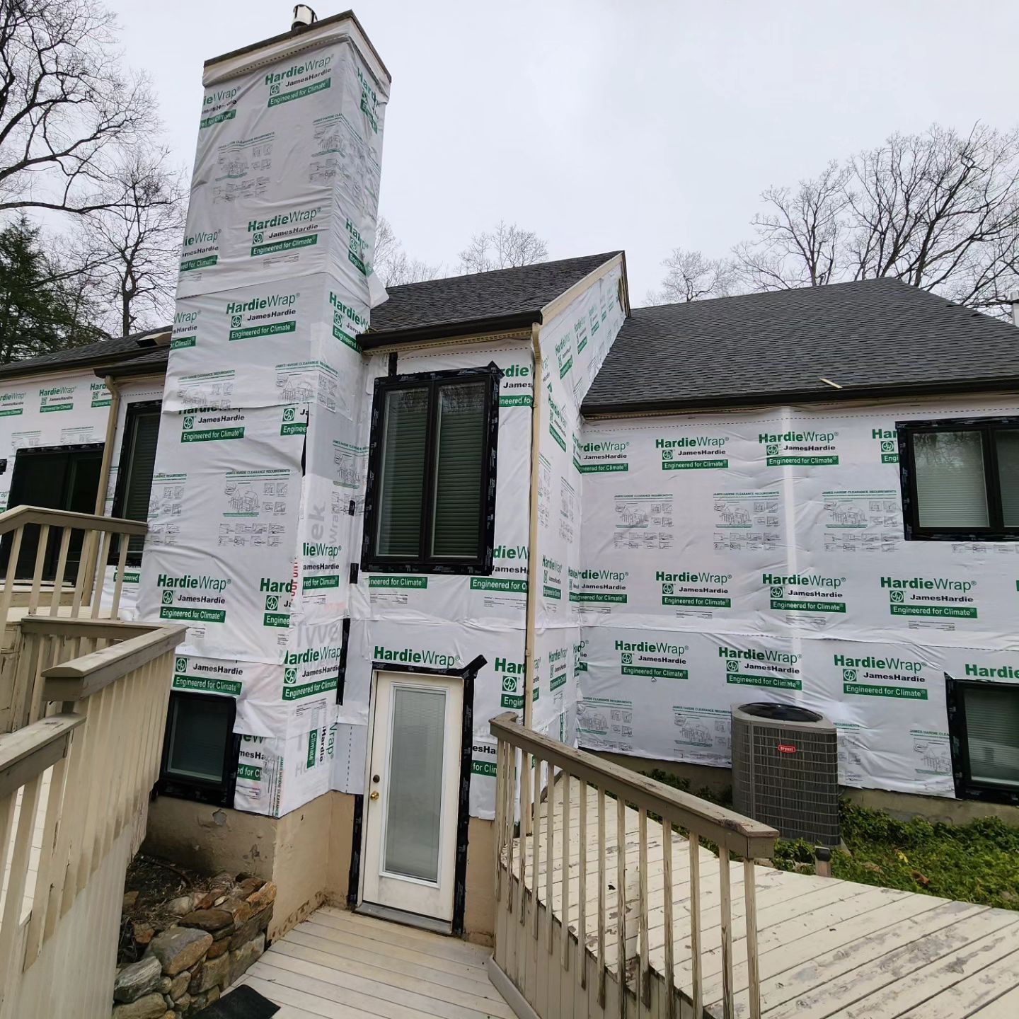 Exterior of a house under construction; white protective wrap covers the walls; chimney and black-framed windows are visible.