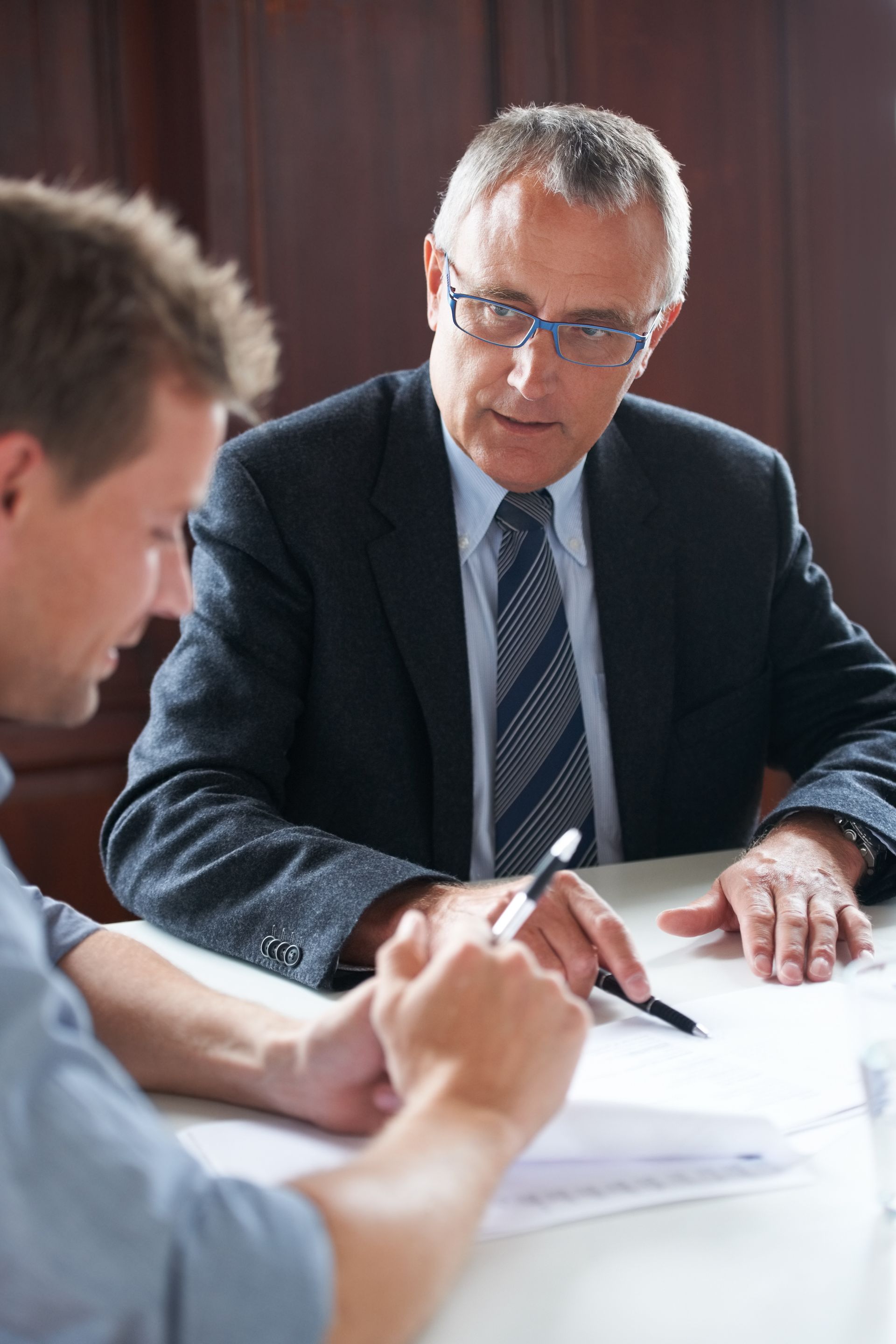 Two men in suits discussing documents at a table, with one speaking and gesturing in a meeting room.