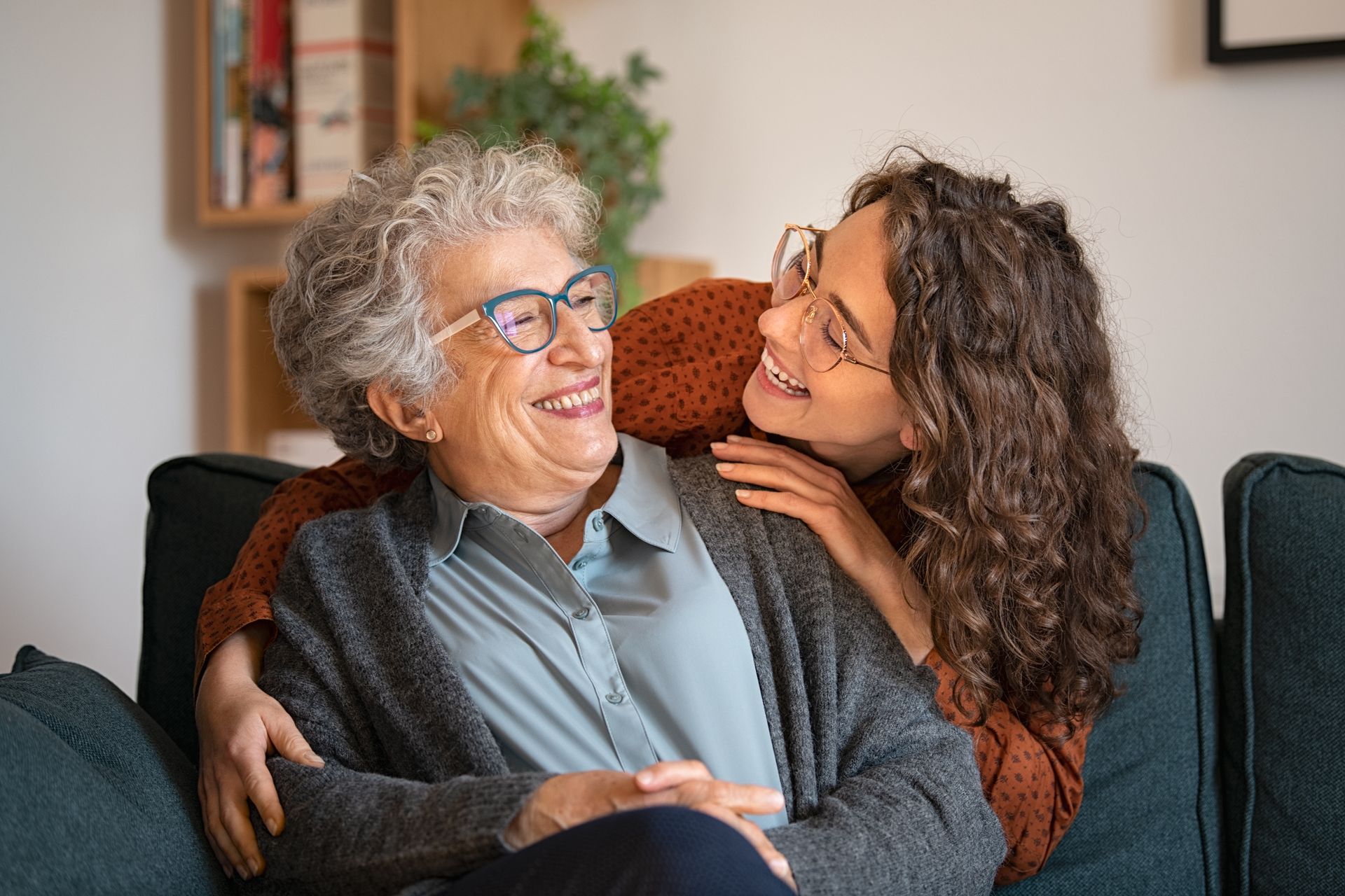 Two people smiling on a couch, one hugging the other in a cozy living room.