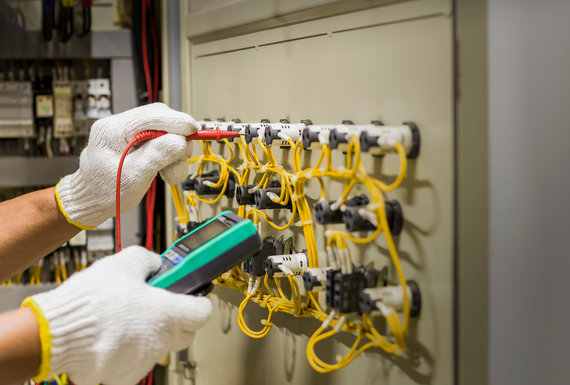 Gloved hands inspect yellow network cables in an electrical panel with a handheld device