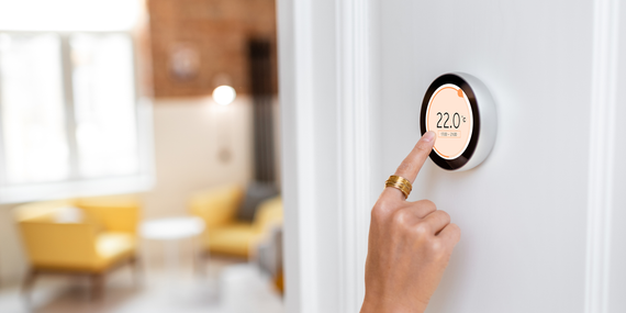Hand holding a doorbell on a white wall, with a bright living room in the background