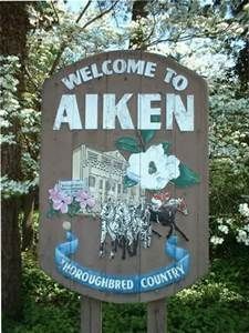 A welcome to aiken sign is surrounded by trees and flowers.