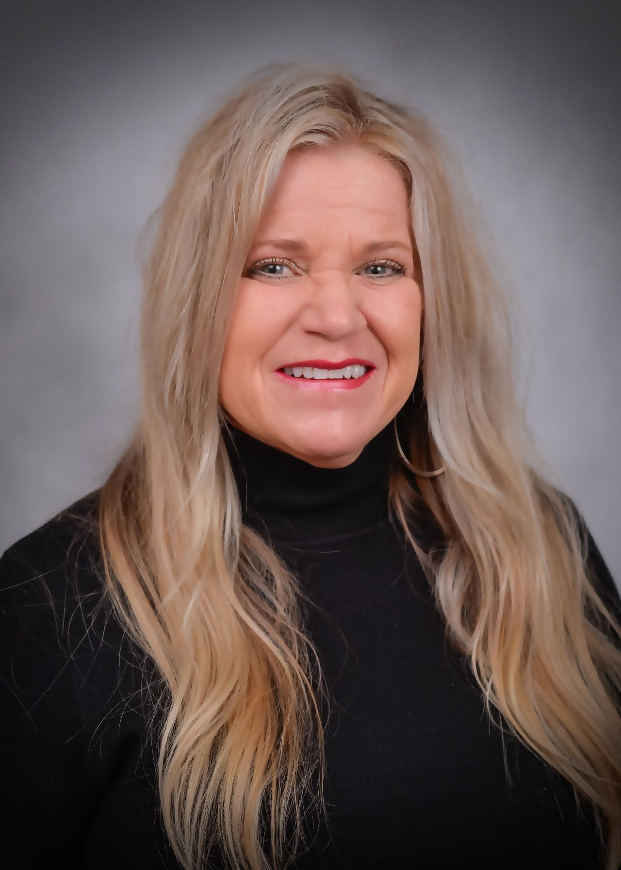 A woman with long blonde hair is wearing a black turtleneck and smiling.