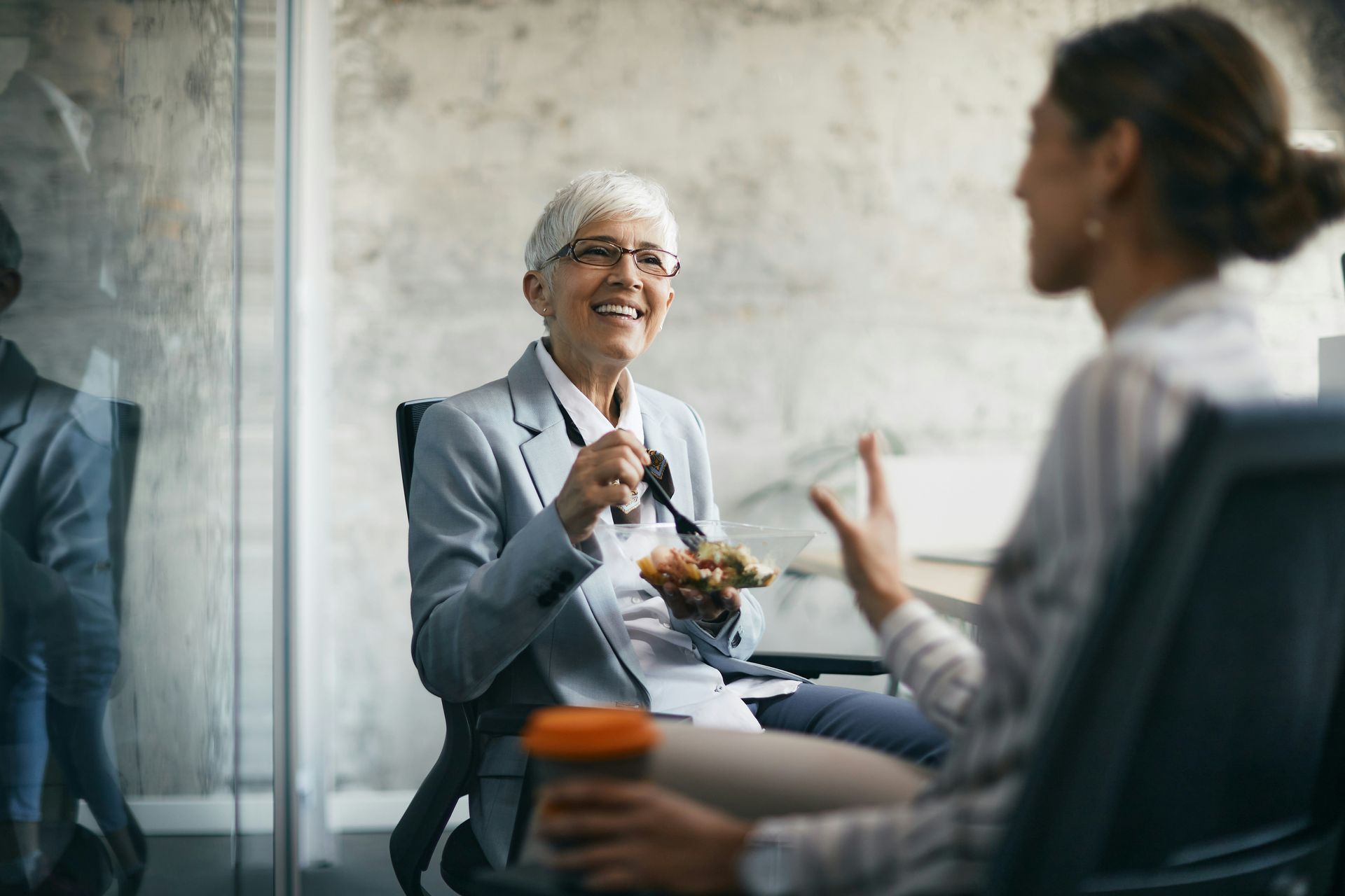 A woman is sitting in a chair talking to another woman while eating a sandwich.