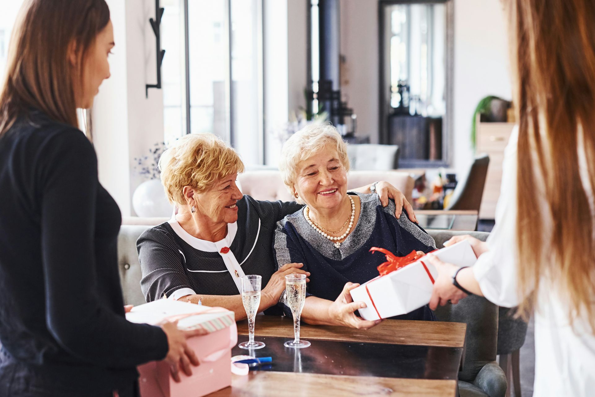 A woman is giving a gift to two older women sitting at a table.